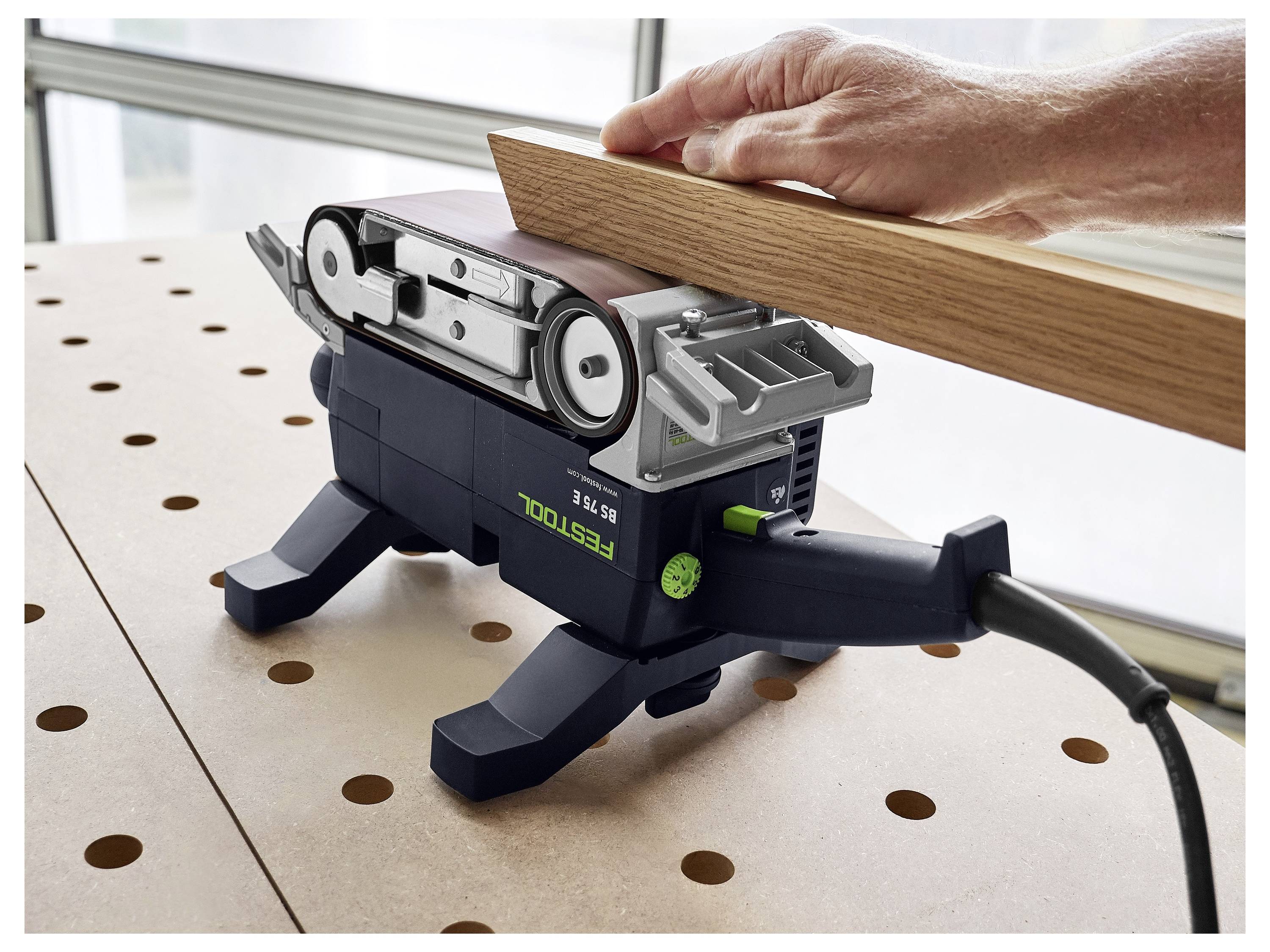 Une personne ponce une planche en bois à l'aide d'une ponceuse à bande électrique sur un établi perforé dans un atelier.