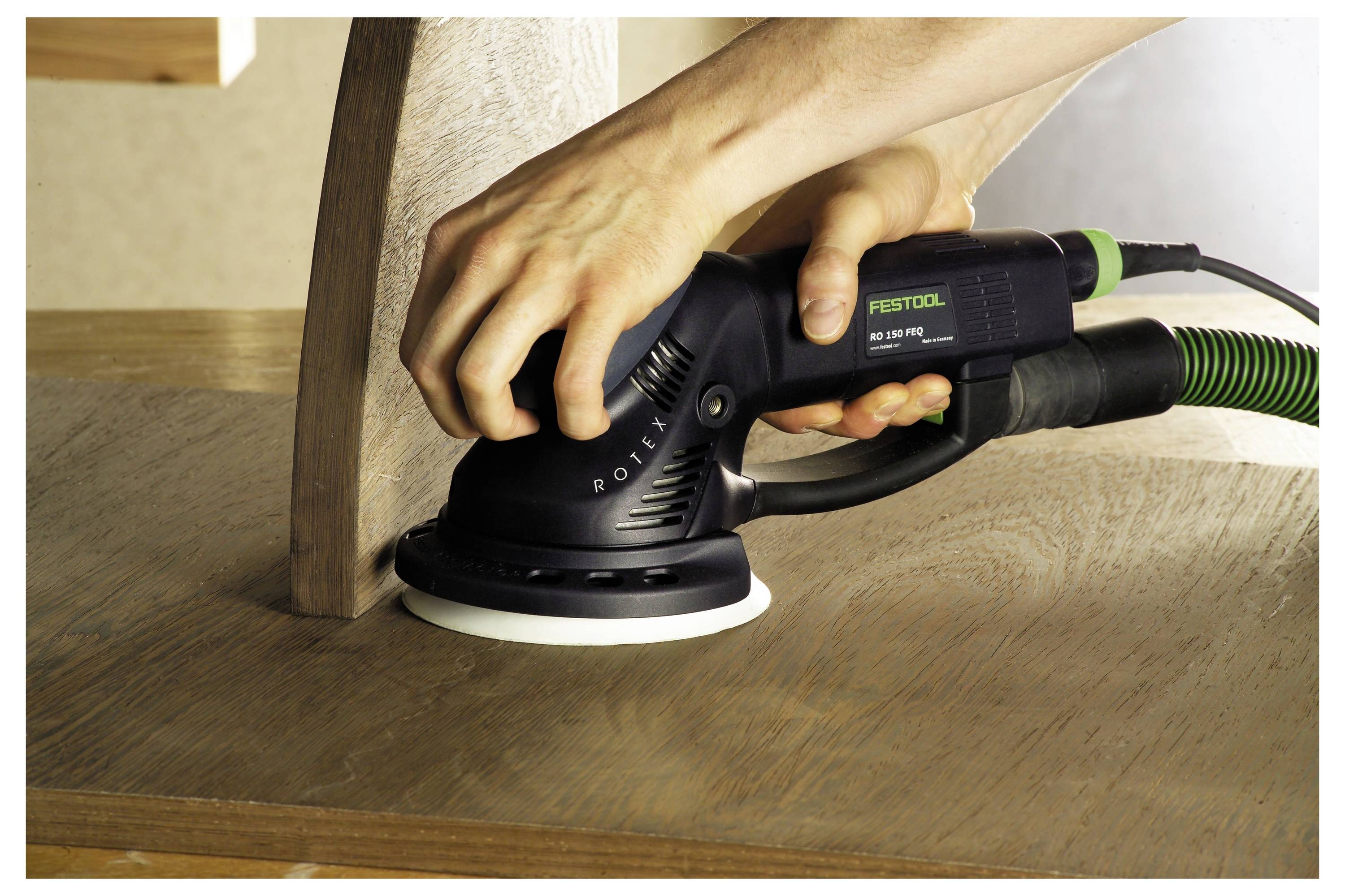 Une personne utilise une ponceuse électrique sur une surface en bois, lissant les bords pour obtenir une finition uniforme. La ponceuse est reliée à un tuyau vert.