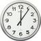 Une horloge murale ronde affichant l'heure 12h03, avec des chiffres noirs et des aiguilles noires sur un cadran blanc, entourée d'un cadre argenté.