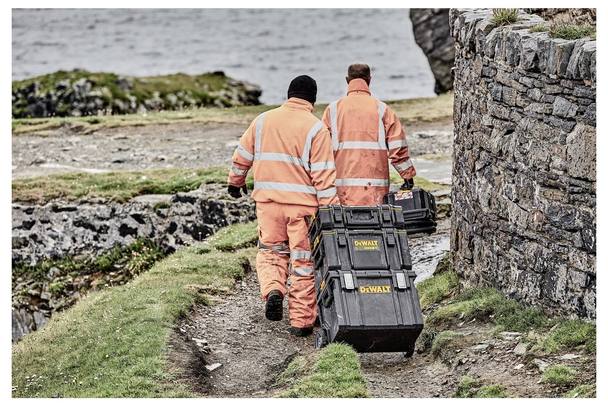 Deux ouvriers vêtus d'équipements de sécurité orange marchent le long d'un chemin près de la mer, tirant de grandes caisses à outils noires. Un solide mur de pierre se trouve au premier plan.