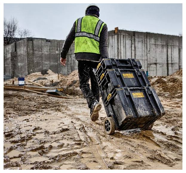 Un ouvrier du bâtiment vêtu d'un gilet de sécurité vert tire une caisse à outils à roulettes sur un chantier de construction boueux avec des murs en béton en arrière-plan.