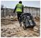 Un ouvrier du bâtiment vêtu d'un gilet de sécurité vert tire une caisse à outils à roulettes sur un chantier de construction boueux avec des murs en béton en arrière-plan.