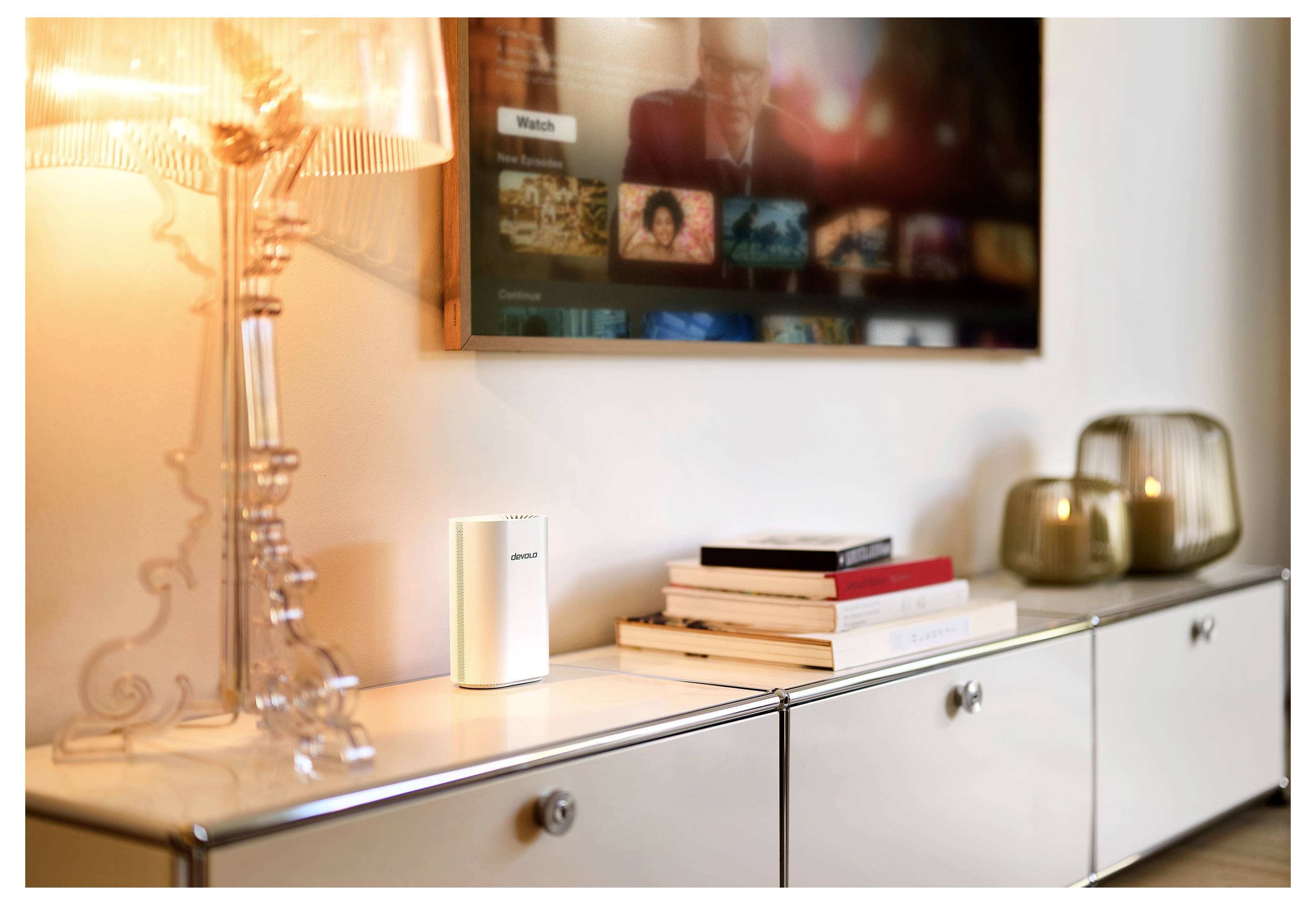 Un salon moderne avec une lampe allumée sur un meuble blanc, des livres empilés, des bougies et un téléviseur affichant divers choix multimédias.