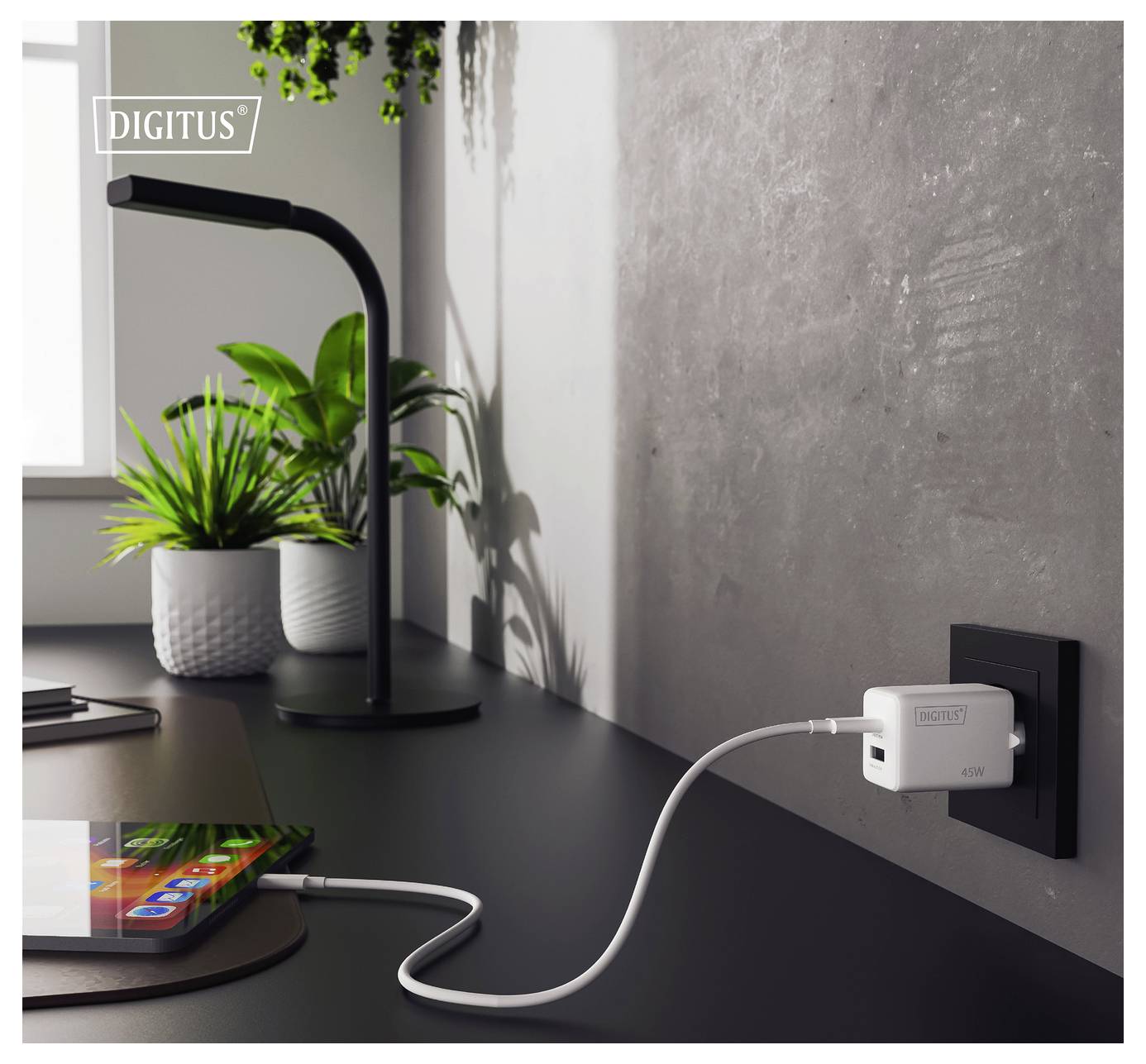 Un smartphone repose sur un bureau connecté à une prise murale via un chargeur USB blanc. Une lampe noire et une plante en pot se trouvent en arrière-plan.