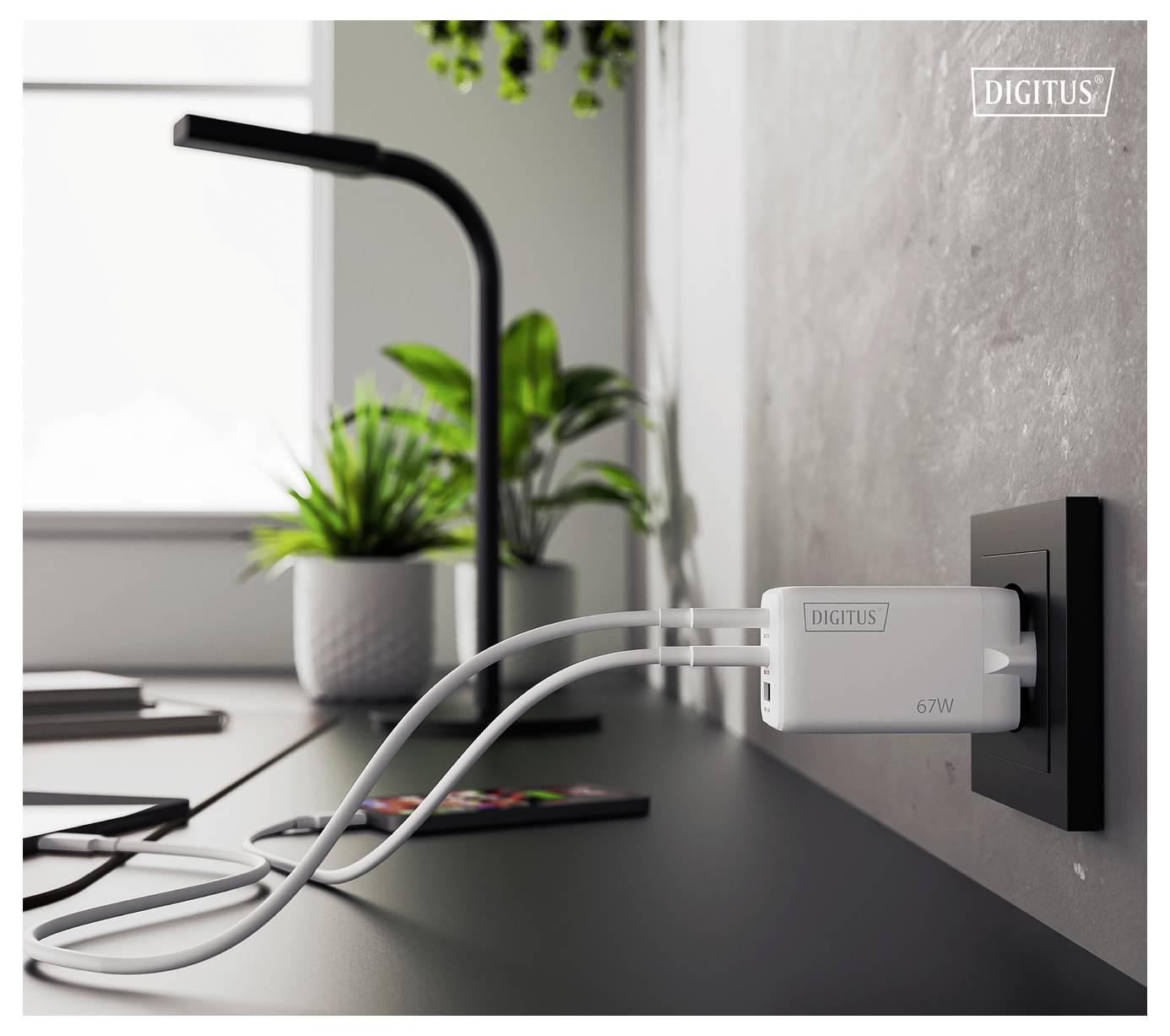 Un bureau minimaliste avec des plantes en pot et une lampe. Un chargeur multi-ports étiqueté « 67W » est branché à une prise murale, alimentant deux appareils.