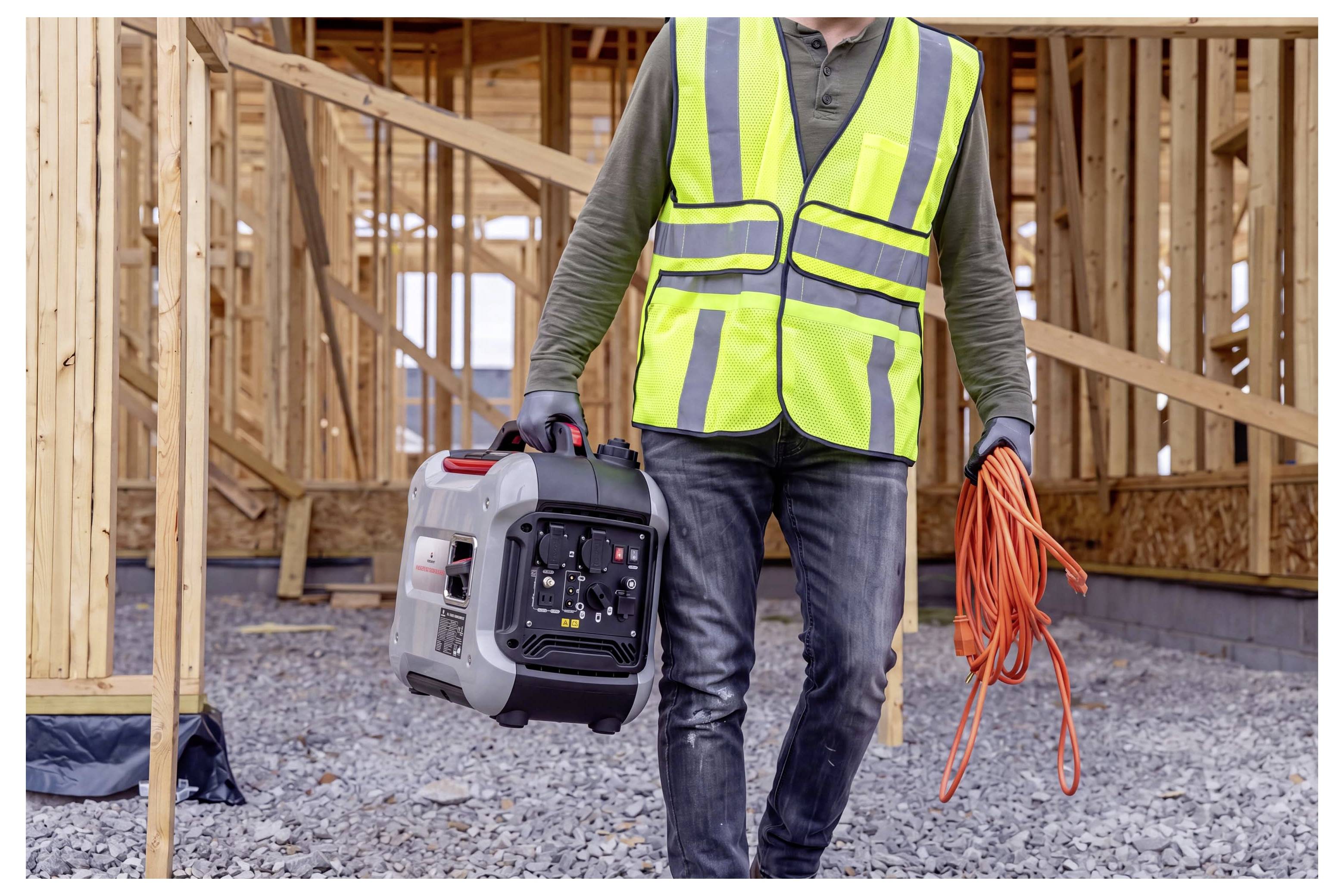 Ouvrier de la construction dans un gilet réfléchissant portant un générateur portable et un câble d'extension orange devant une maison en construction.