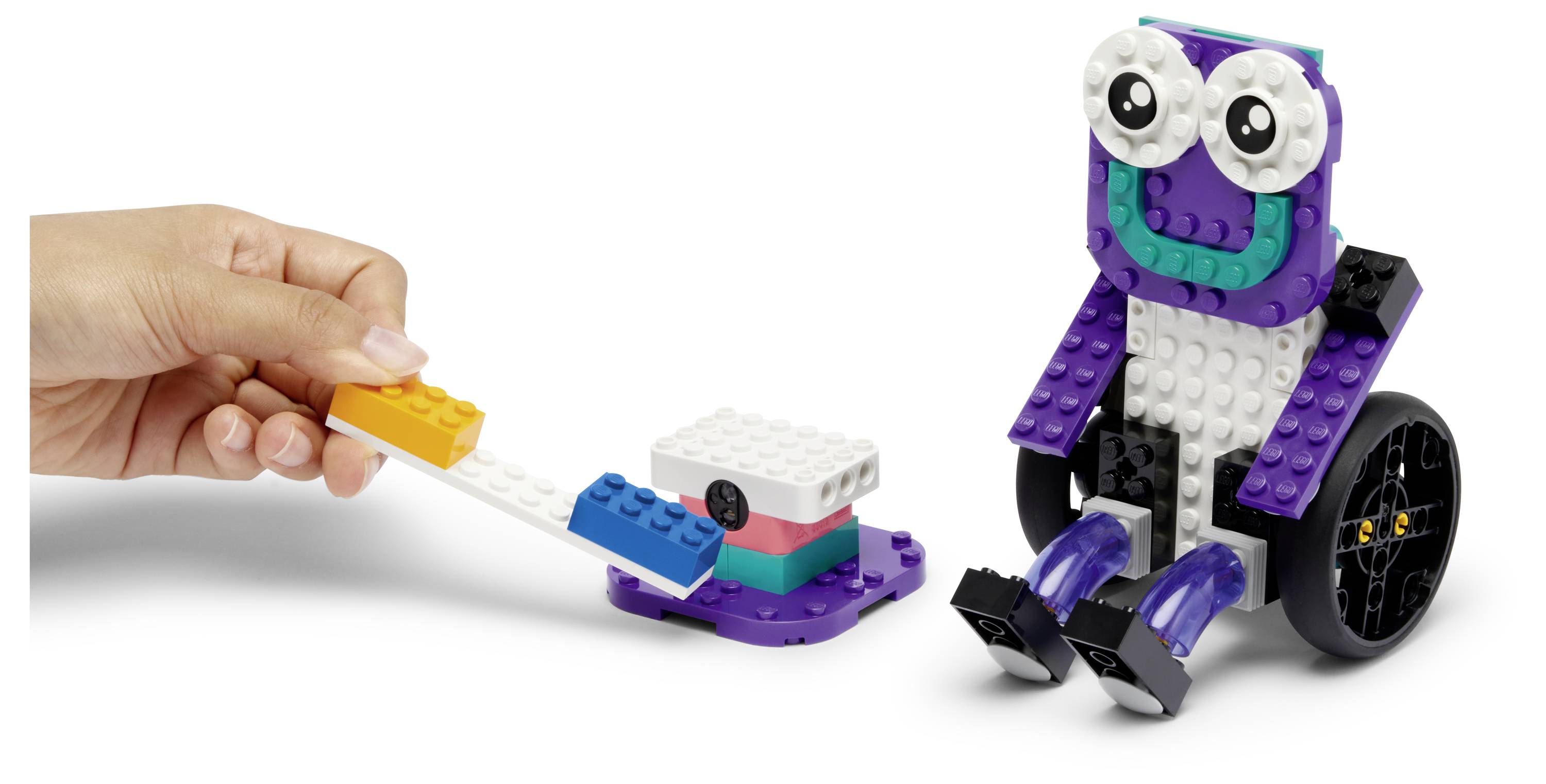 Une main interagit avec un petit robot LEGO aux grands yeux et aux roues, démontrant la programmation avec un lecteur de code en briques colorées.