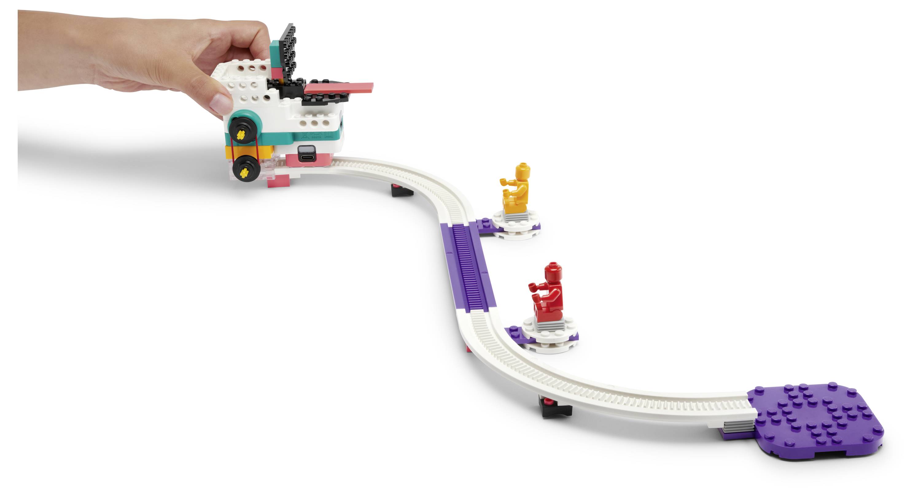 Une main place un petit jouet coloré sur une piste surélevée et courbe dotée de deux figurines jouets.