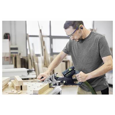 Homme portant des lunettes de sécurité utilisant une biscuiteuse Festool sur une planche de bois dans un atelier. Des pièces de bois et des outils sont visibles en arrière-plan.