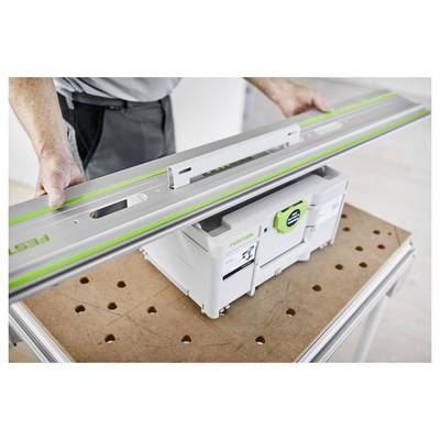 Personne tenant un long outil électrique plat au-dessus d'un boîtier de rangement Festool vert et blanc sur un établi perforé.