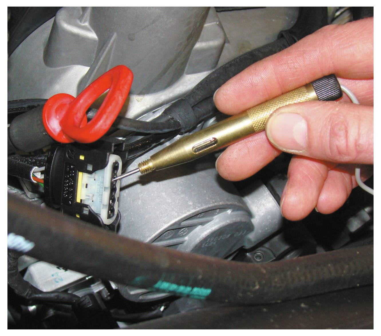 Une main utilise un testeur de tension pour vérifier un connecteur électrique dans une voiture. La sonde touche une broche, avec des fils visibles en arrière-plan.