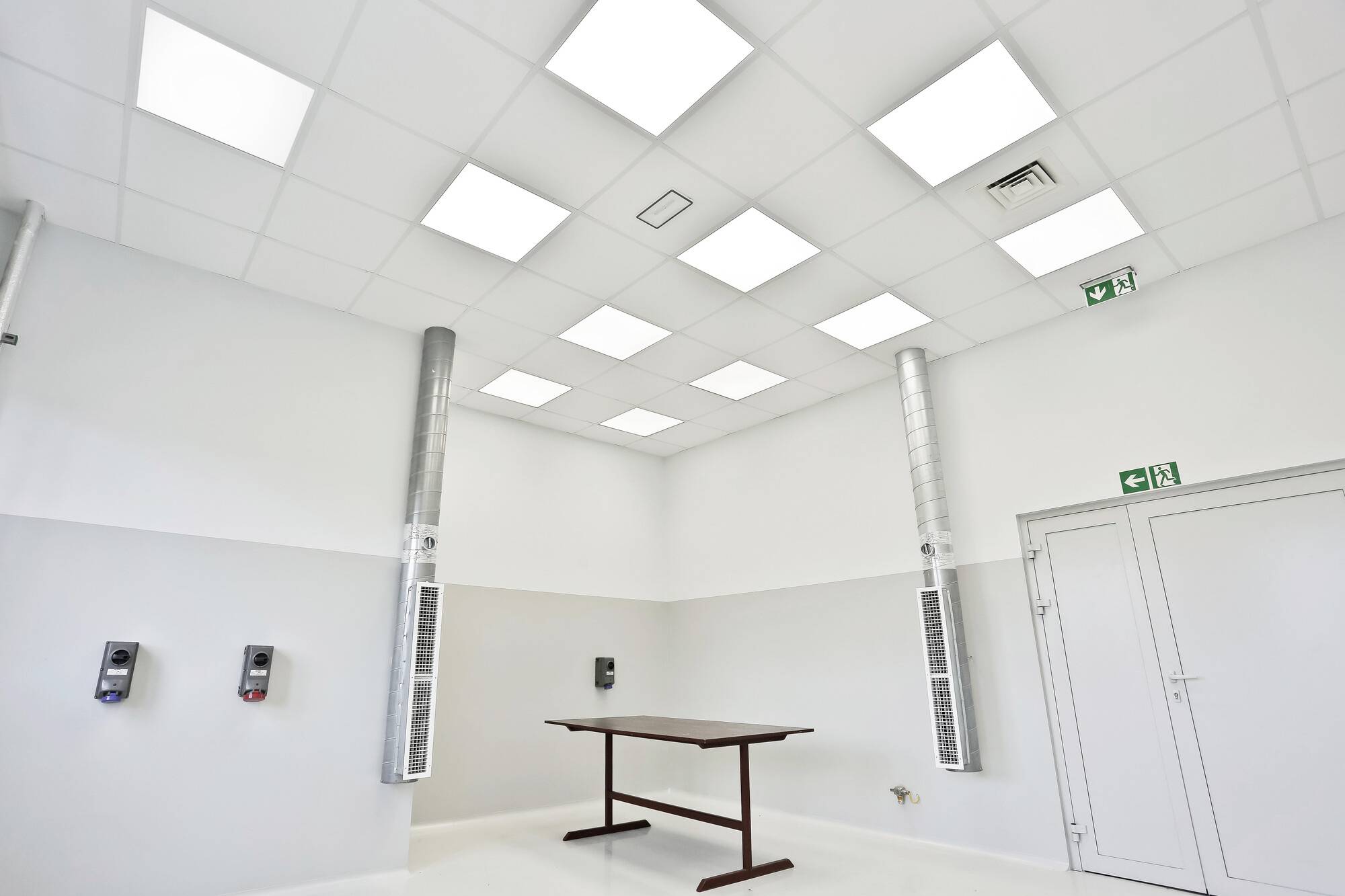 Une pièce avec un plafond haut dotée de luminaires LED carrés au plafond, deux tubes de ventilation, trois appareils muraux et une table marron.