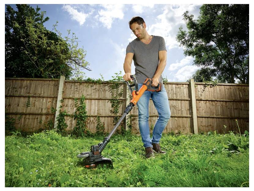 Une personne utilise un coupe-herbe électrique orange et noir pour tondre le gazon dans une cour arrière avec une clôture en bois et des arbres en arrière-plan.