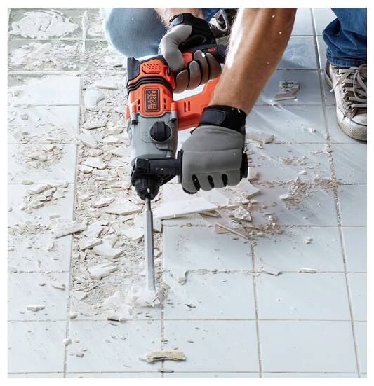 Une personne utilise un outil électrique pour enlever des carreaux d'un sol, ce qui entraîne des morceaux cassés dispersés autour. L'outil est tenu avec des gants.