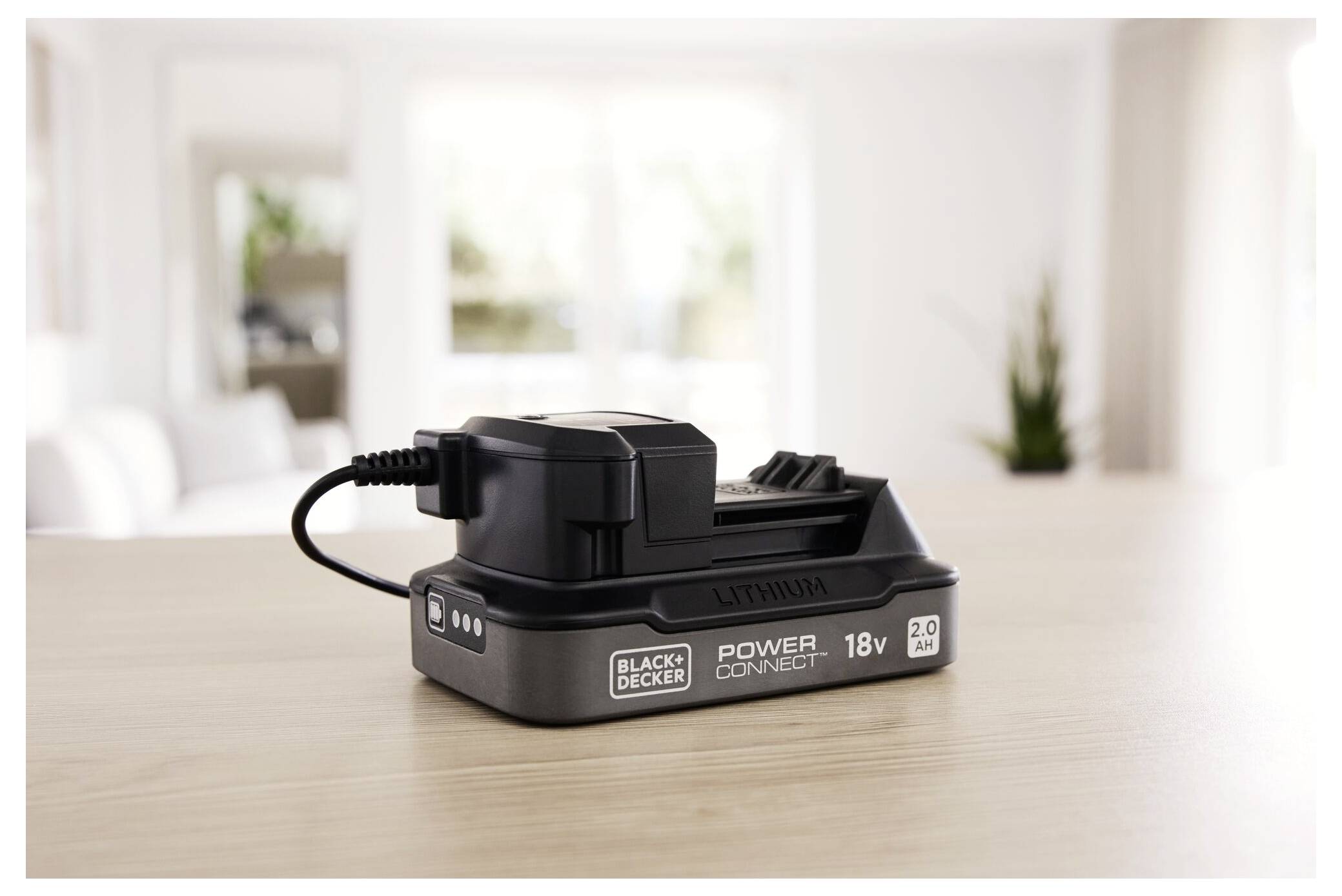 Une batterie au lithium « Black+Decker Power Connect 18V 2.0 Ah » est posée sur une table en bois dans un salon moderne et lumineux.