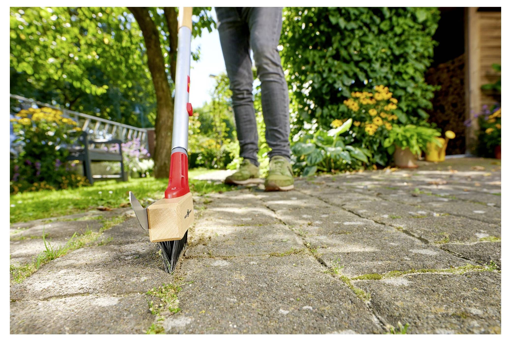 'Personne utilisant un désherbeur pour enlever les mauvaises herbes entre les pierres de patio dans un jardin avec des fleurs et de la verdure en arrière-plan.'