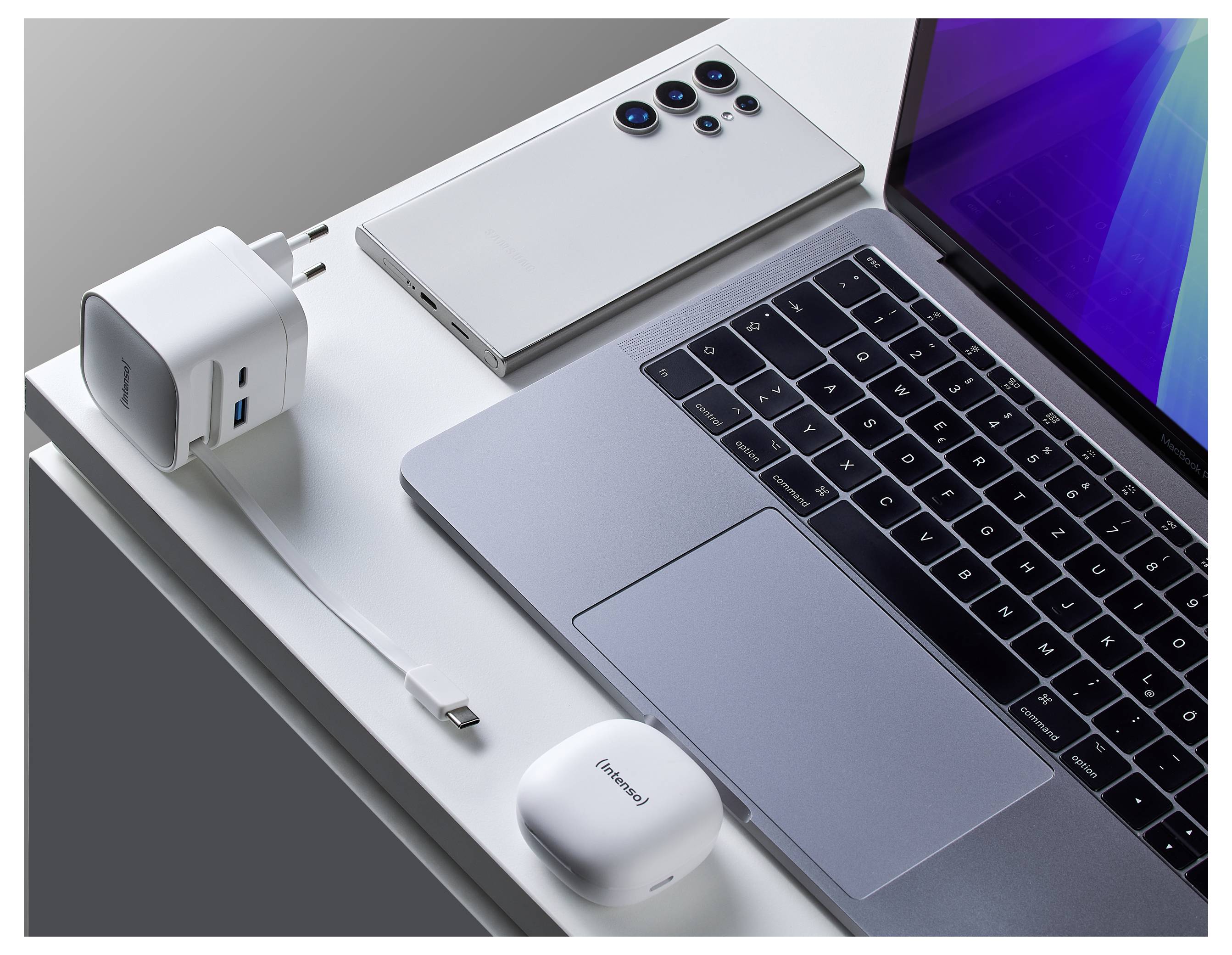 Un ordinateur portable sur un bureau avec un smartphone, un chargeur double port et un étui blanc pour écouteurs sans fil.