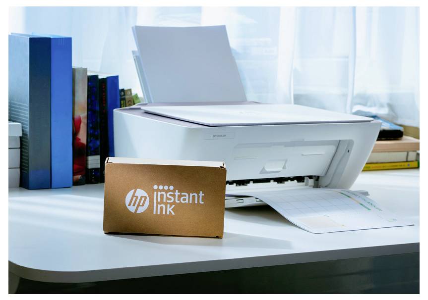 Une imprimante blanche sur un bureau, à côté de livres, avec une boîte HP Instant Ink au premier plan. La lumière filtre par une fenêtre derrière.