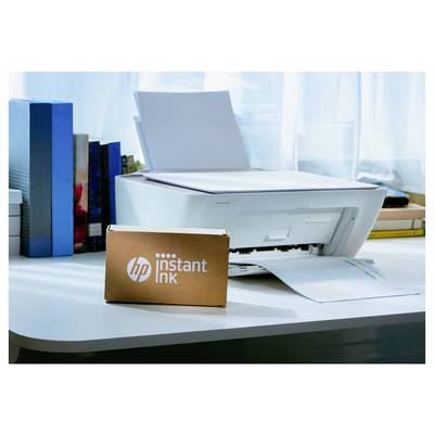 Une imprimante blanche sur un bureau, à côté de livres, avec une boîte HP Instant Ink au premier plan. La lumière filtre par une fenêtre derrière.