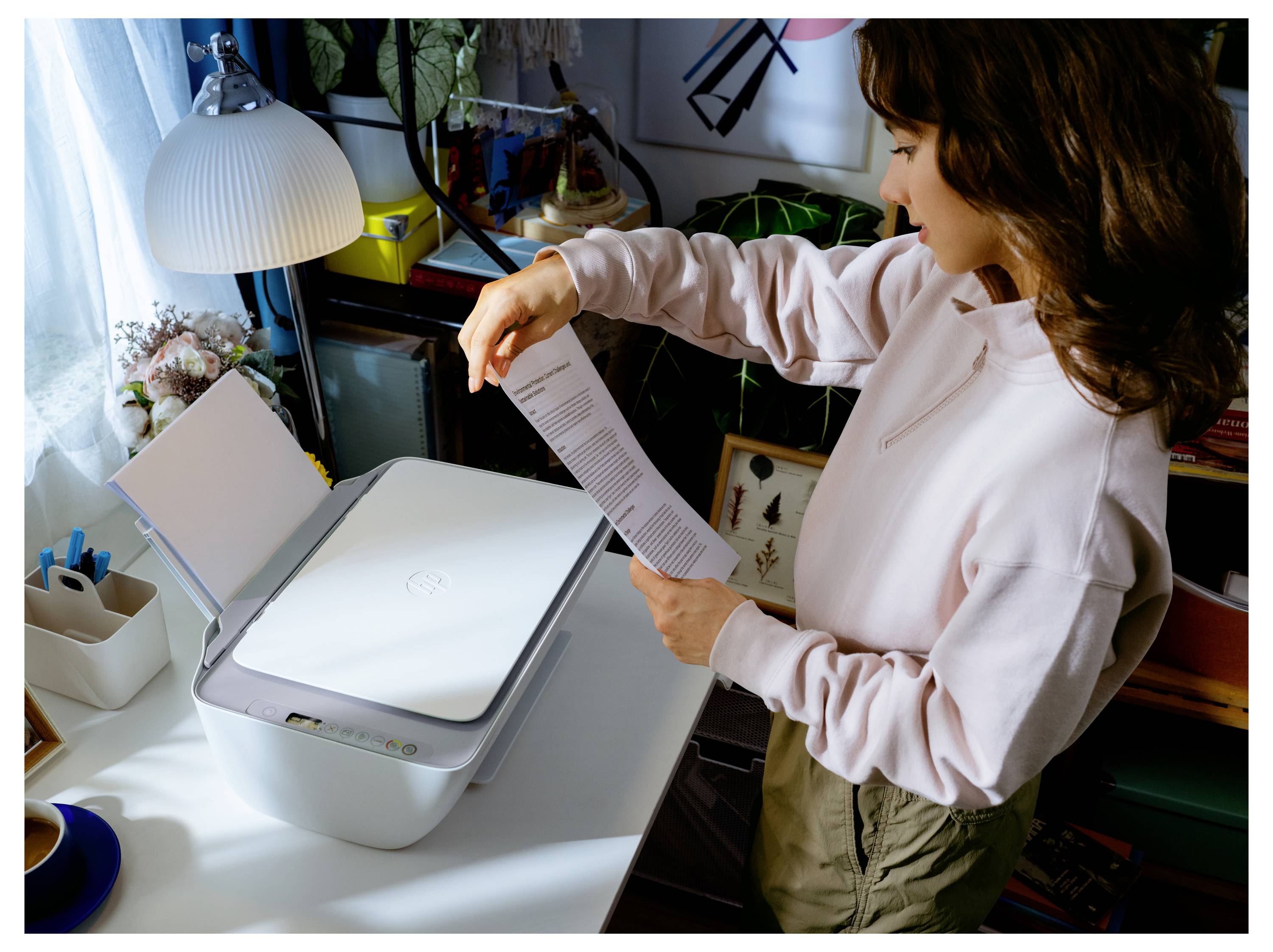 Une femme dans un bureau à domicile charge un document dans un scanner à plat situé à côté d'un ordinateur portable et d'une lampe de bureau.