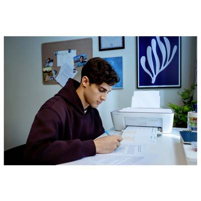 Une personne vêtue d'un sweat-shirt à capuche est assise à un bureau et écrit sur des papiers avec une imprimante à proximité. Le mur affiche un tableau d'affichage et des œuvres d'art encadrées.