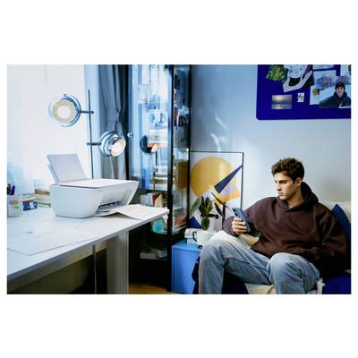Une personne est assise sur un canapé en regardant un smartphone, à côté d'une table avec une imprimante et des papiers. Un meuble avec des livres et des objets de décoration artistique se trouve à proximité.