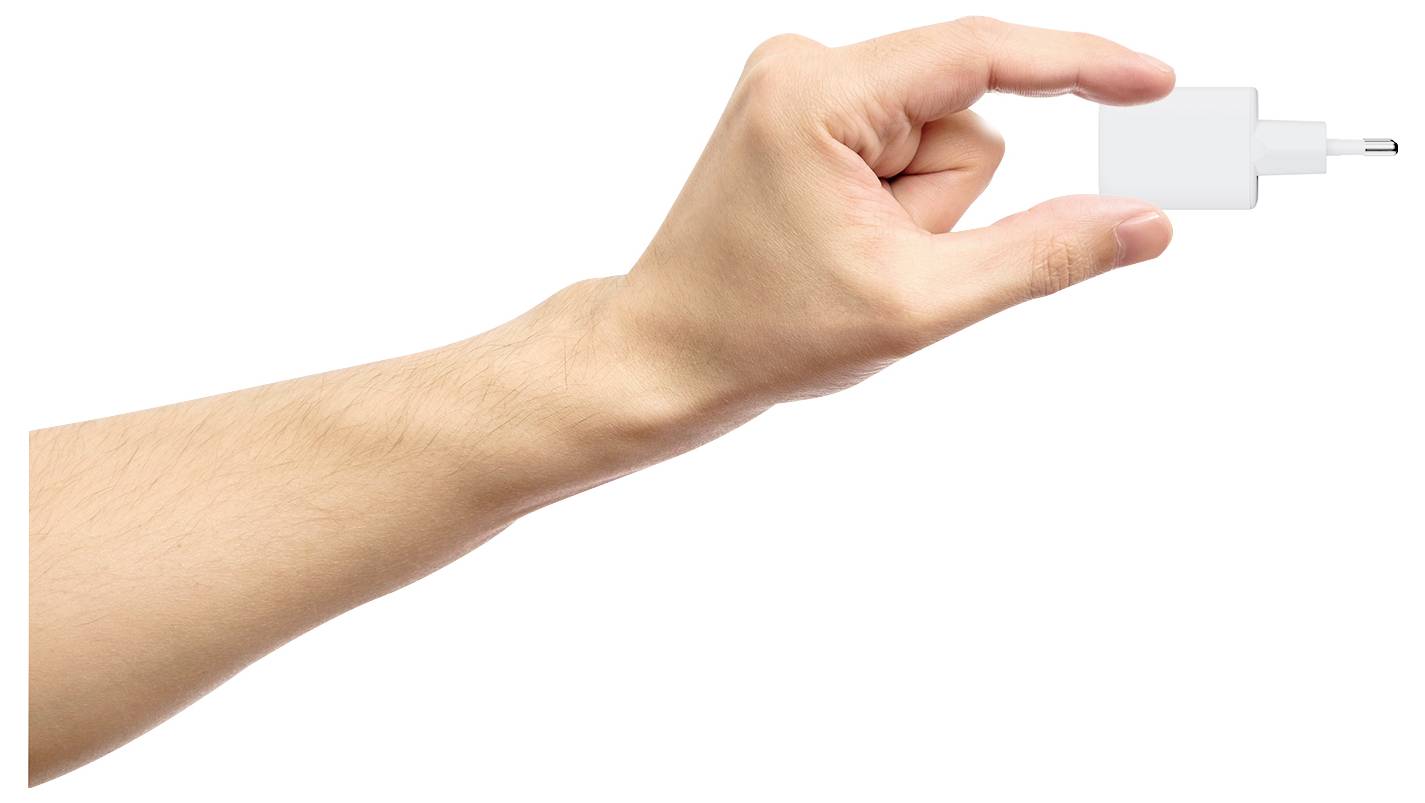 Une main tenant une petite prise de chargeur USB blanc pincée entre le pouce et l'index, sur un fond blanc uni.