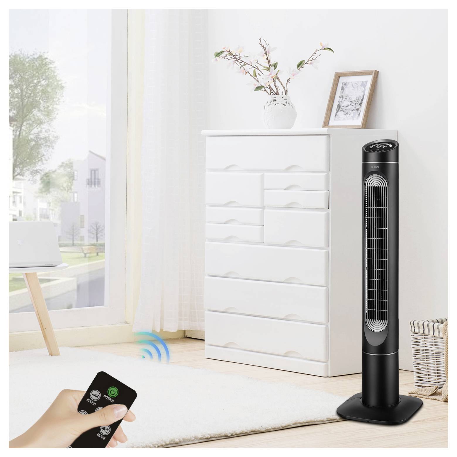 Une personne utilise une télécommande pour contrôler un grand ventilateur de tour noir dans une pièce lumineuse avec une commode blanche, une plante et une photo encadrée sur le dessus.