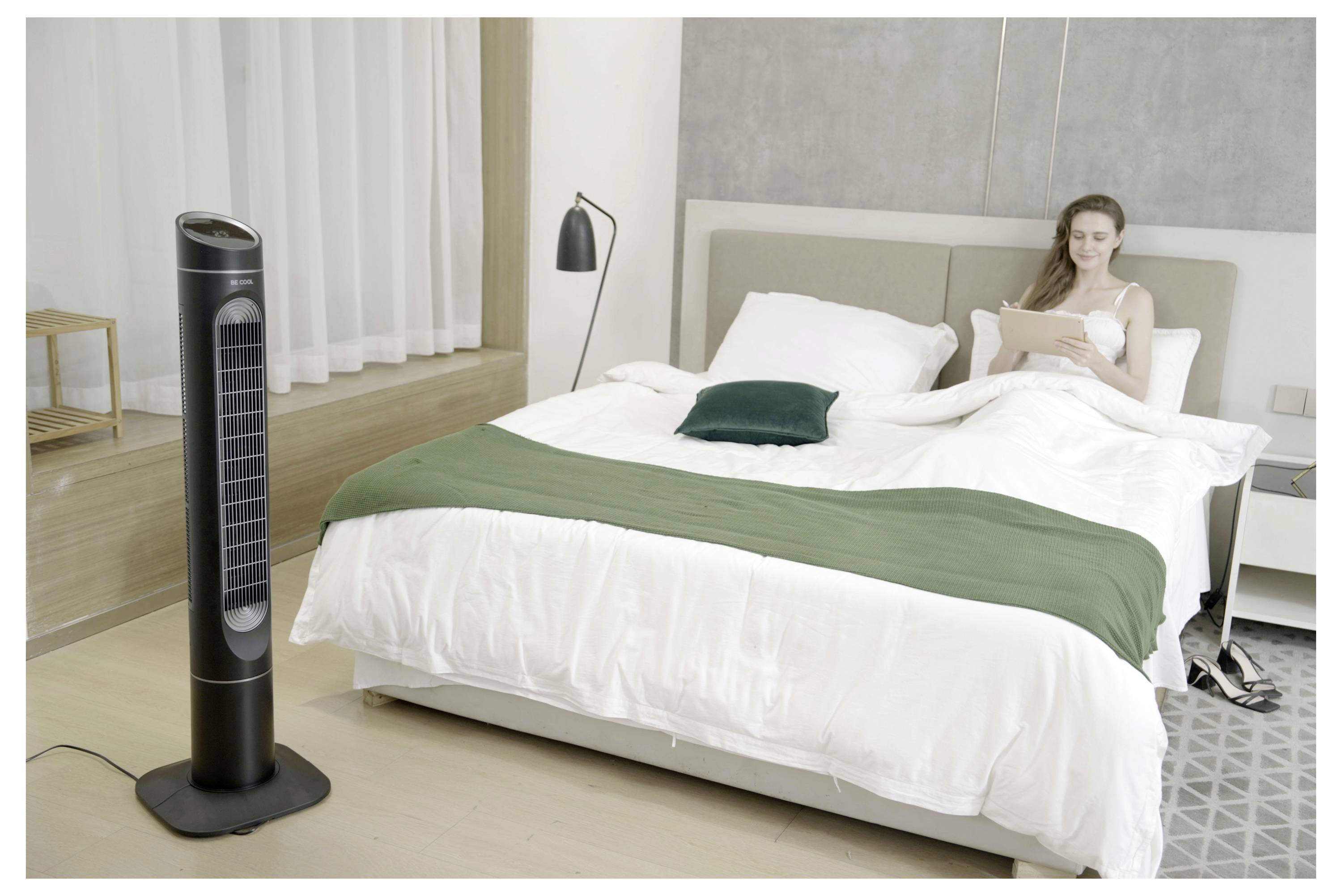 Une femme est assise sur un lit dans une chambre à coucher moderne et lumineuse, lisant une tablette. Un grand ventilateur noir se tient à proximité, suggérant une attention portée au confort et à la détente.