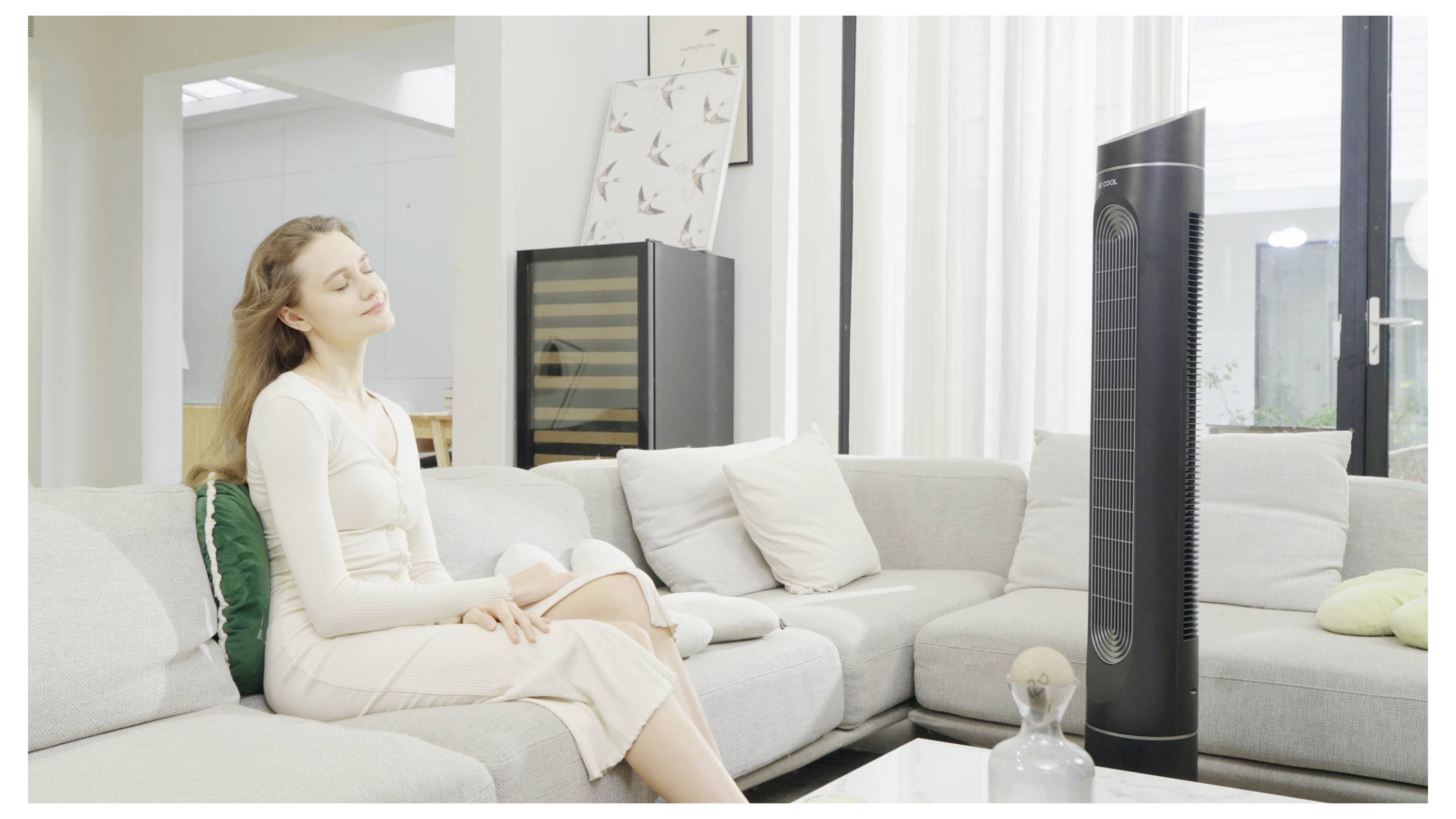 Une femme est assise, détente, sur un canapé, les yeux fermés, profitant de la brise d'un grand ventilateur noir dans un cadre de salon moderne.