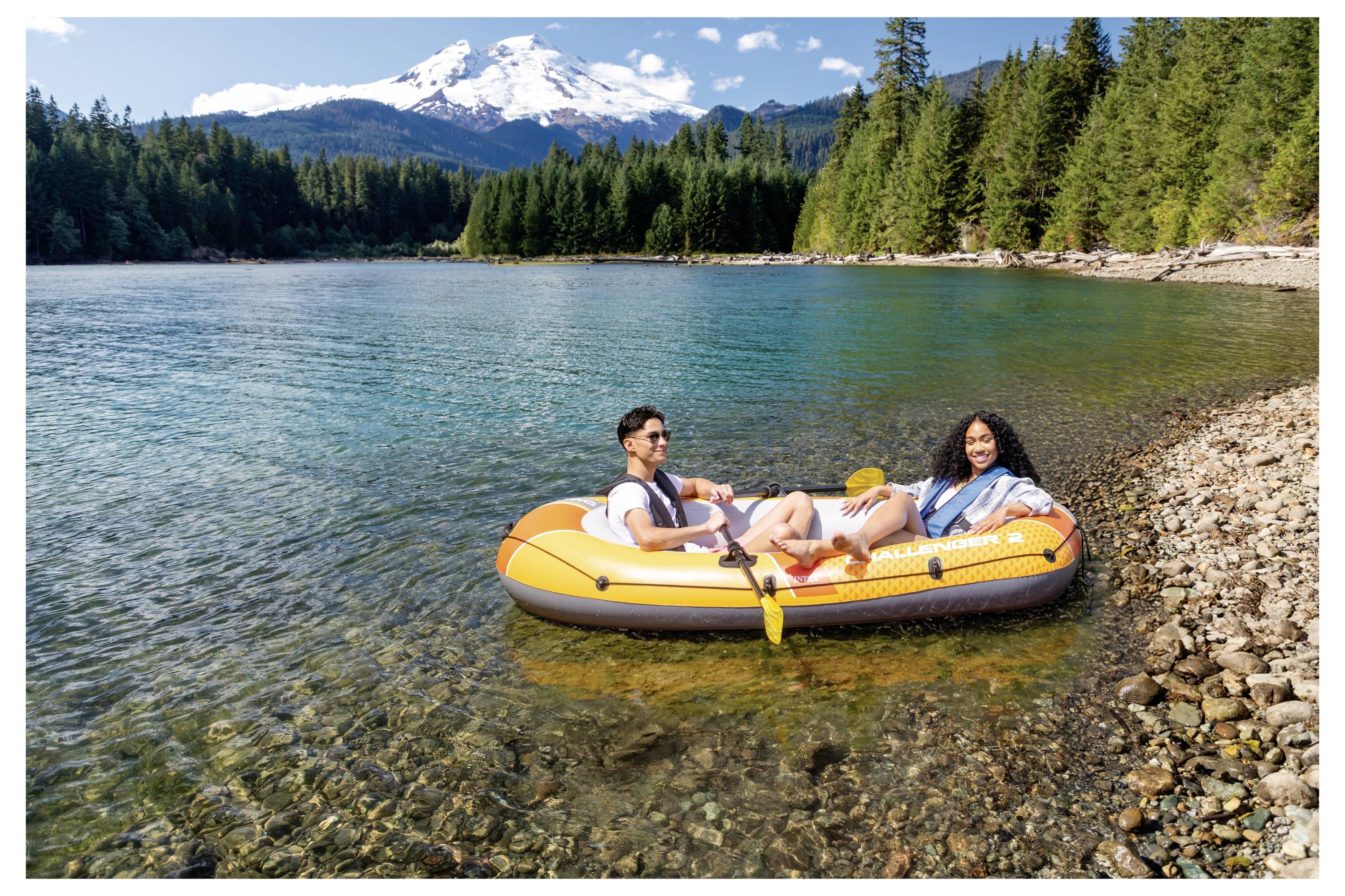 Un homme et une femme se détendent dans un bateau gonflable coloré sur un lac cristallin entouré de forêt, avec une montagne enneigée en arrière-plan.