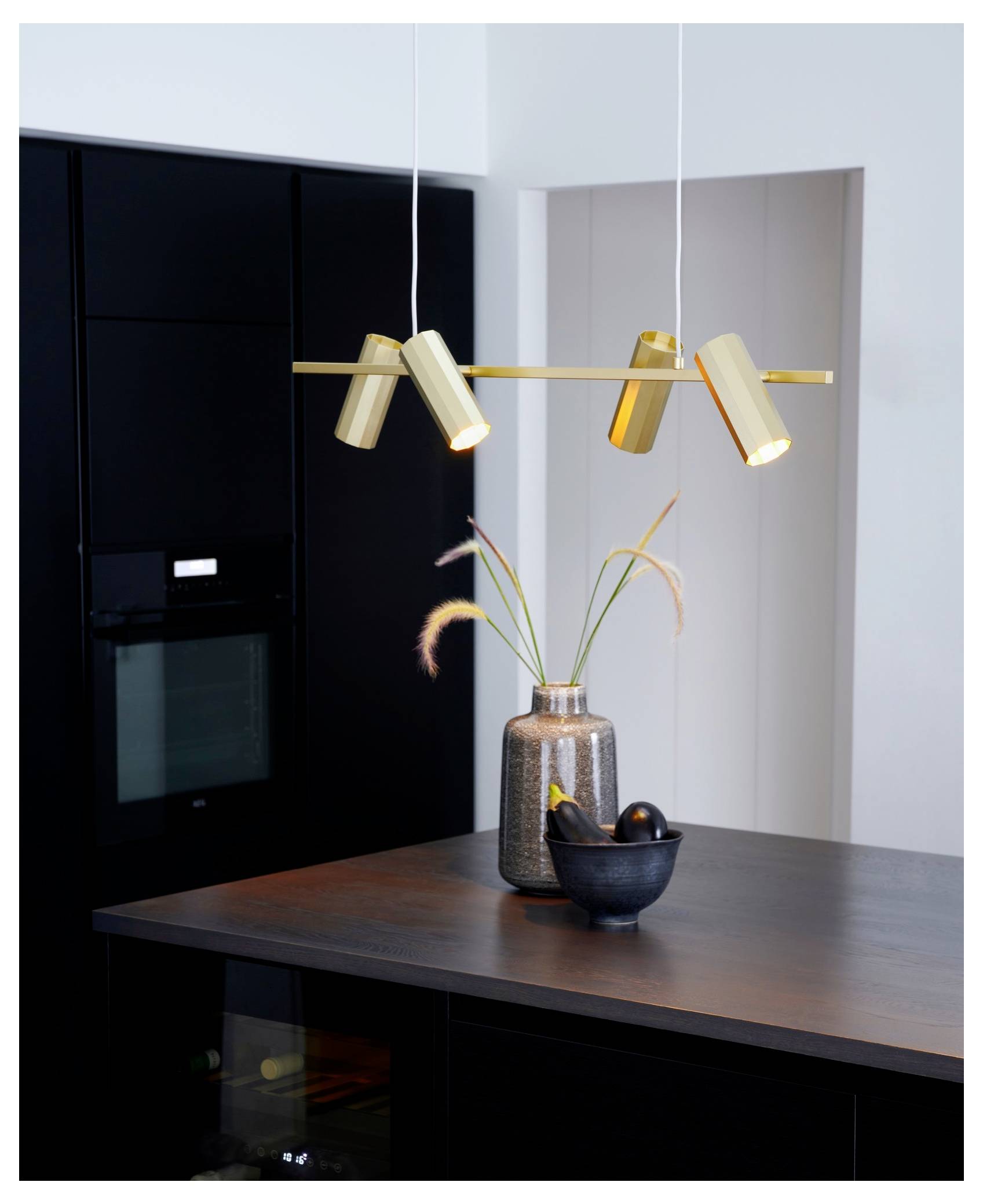Une cuisine moderne avec un four noir épuré et un plan de travail sophistiqué est éclairée par une suspension dorée. Un vase décoratif avec des plantes et un bol sont posés sur le plan de travail.