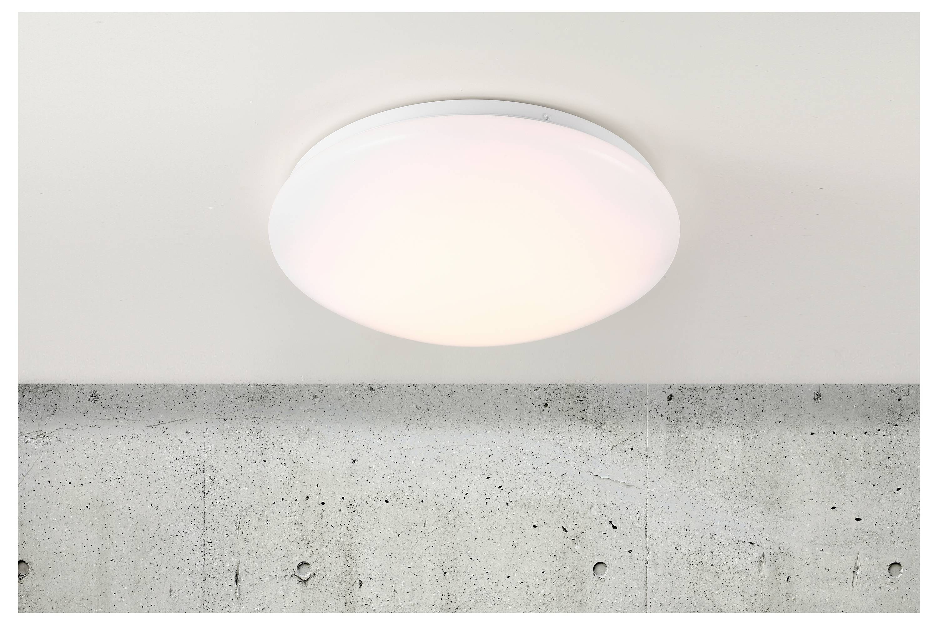 Luminaire de plafond monté sur un plafond blanc, au-dessus d'un mur en béton. Le luminaire émet une lueur blanche douce, indiquant qu'il est allumé.