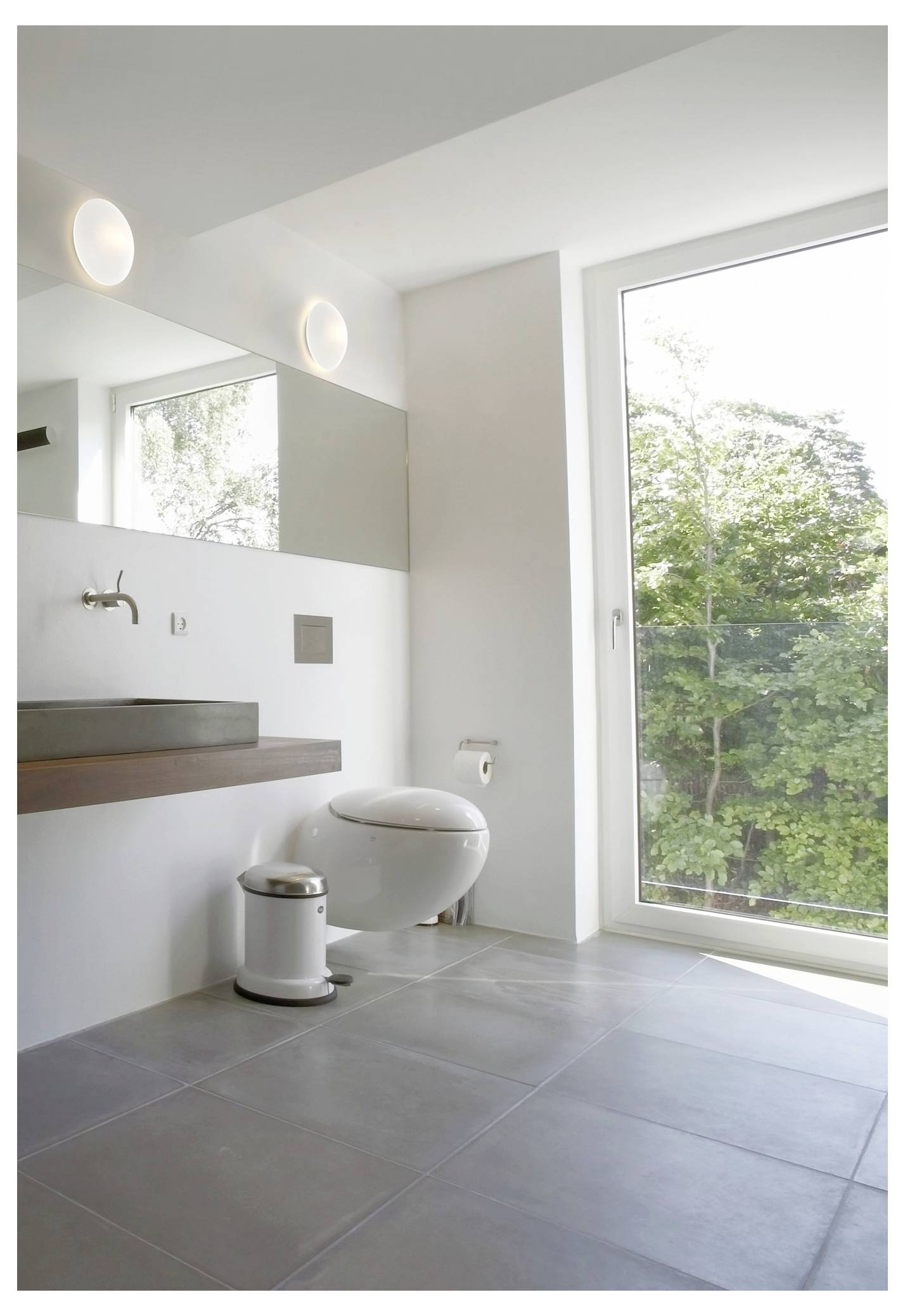 Une salle de bain moderne avec une toilette blanche, un lavabo rectangulaire, une grande fenêtre avec vue sur les arbres, et une décoration minimaliste.
