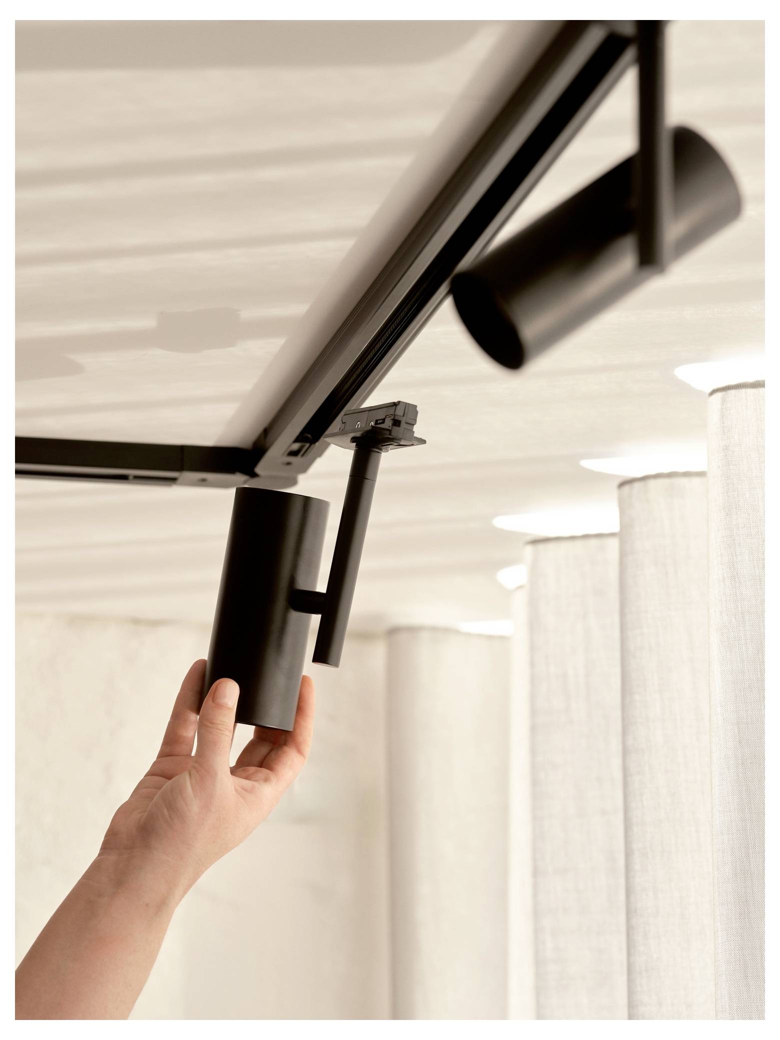 Une main ajuste un luminaire de plafond cylindrique noir dans une pièce faiblement éclairée, près d'une rangée de rideaux blancs.