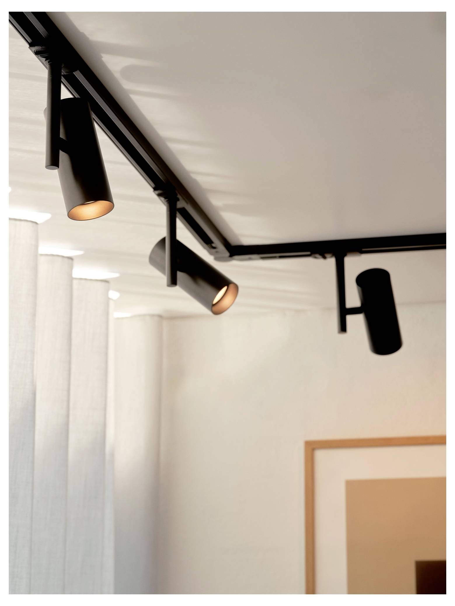 Appareils d'éclairage sur rail fixés à un plafond blanc dans un intérieur moderne, avec un appareil allumé et une œuvre d'art encadrée partiellement visible en dessous.