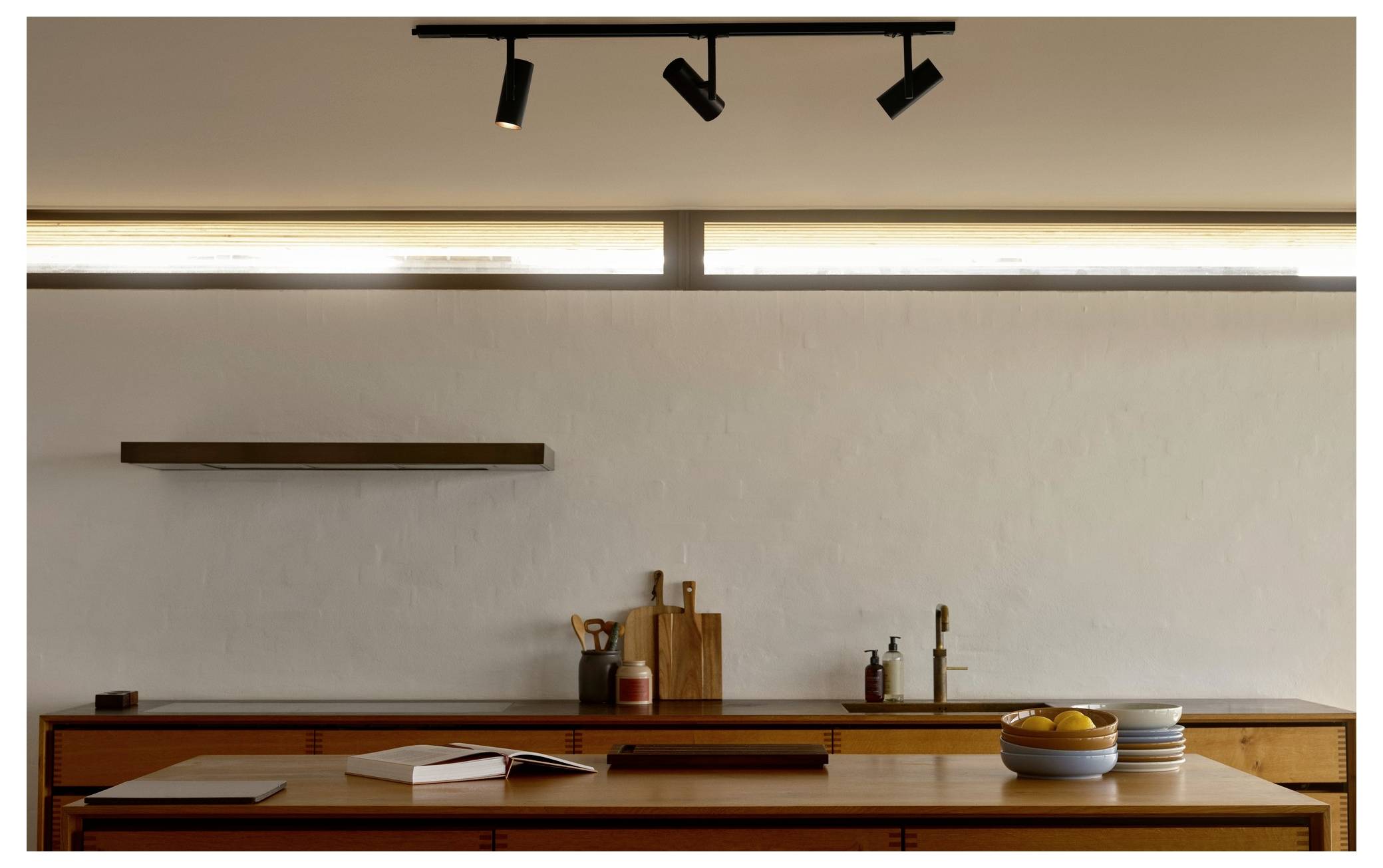 Une cuisine minimaliste avec des plans de travail en bois, des planches à découper et un compotier. Un éclairage subtil provenant de projecteurs encastrés au-dessus illumine l'espace.
