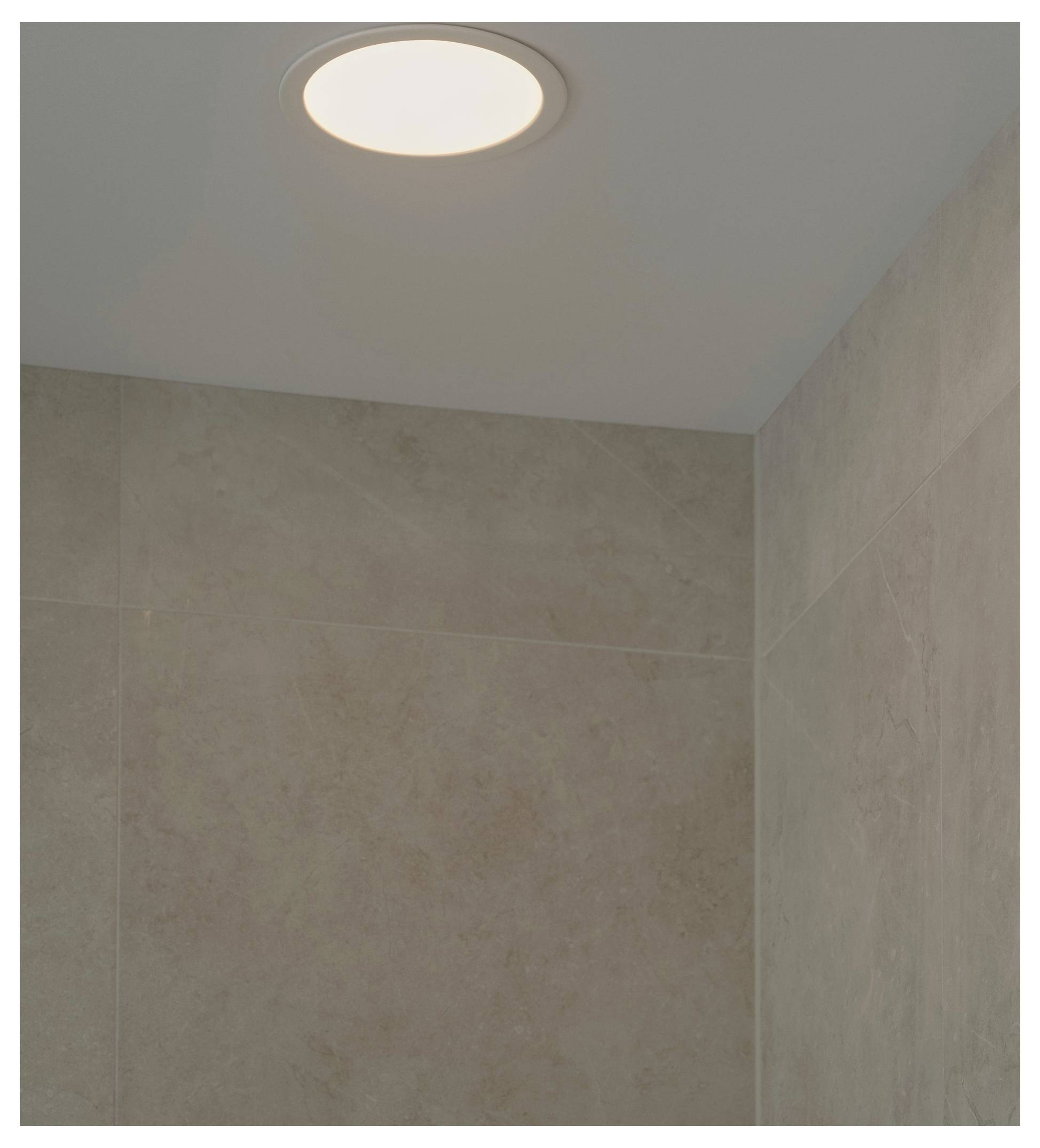 Une lumière de plafond encastrée illumine un coin de la pièce aux murs carrelés beige, mettant en évidence la texture lisse et les tons neutres.