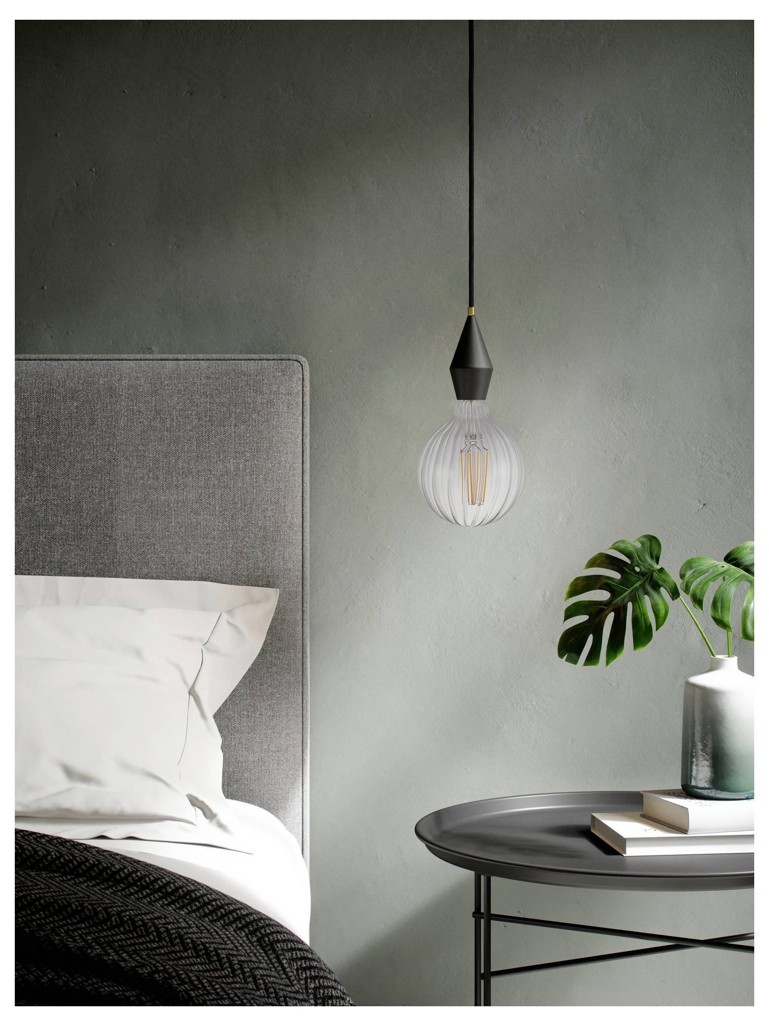 Une chambre moderne avec une tête de lit rembourrée grise, de la literie blanche, une suspension luminaire, et une petite table avec une plante et des livres.