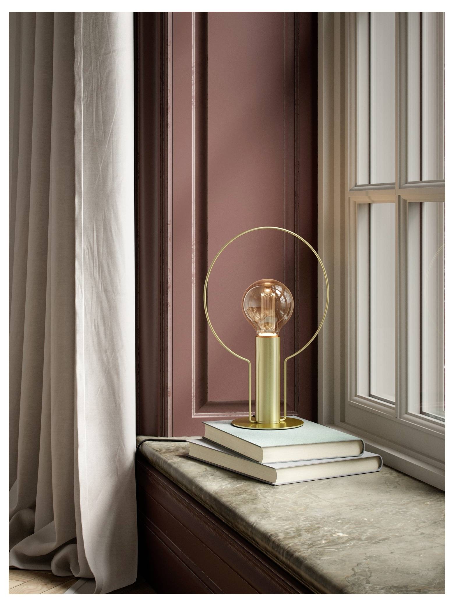 Une lampe moderne avec un cadre métallique circulaire et une ampoule apparente repose sur des livres près d'une fenêtre aux panneaux roses et aux rideaux qui vont du sol au plafond.