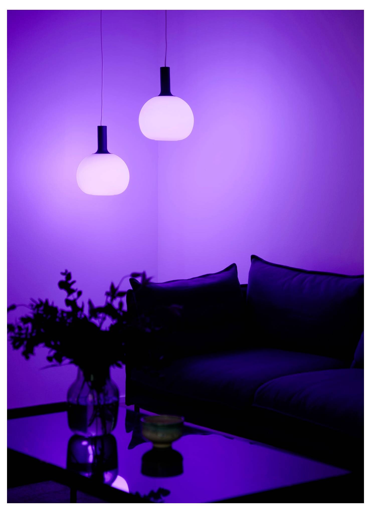 Une pièce faiblement éclairée avec deux lampes suspendues luminescentes et un canapé. Une table en verre avec un vase de fleurs et un petit bol ajoute à l'ambiance chaleureuse.