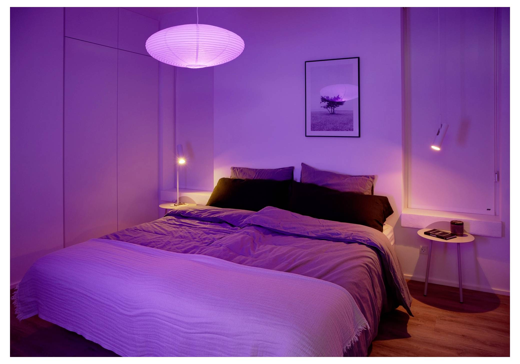 Une chambre à coucher avec un lit double parfaitement fait, un éclairage violet doux, une lampe en papier ronde au-dessus, et une petite table de nuit avec un livre.