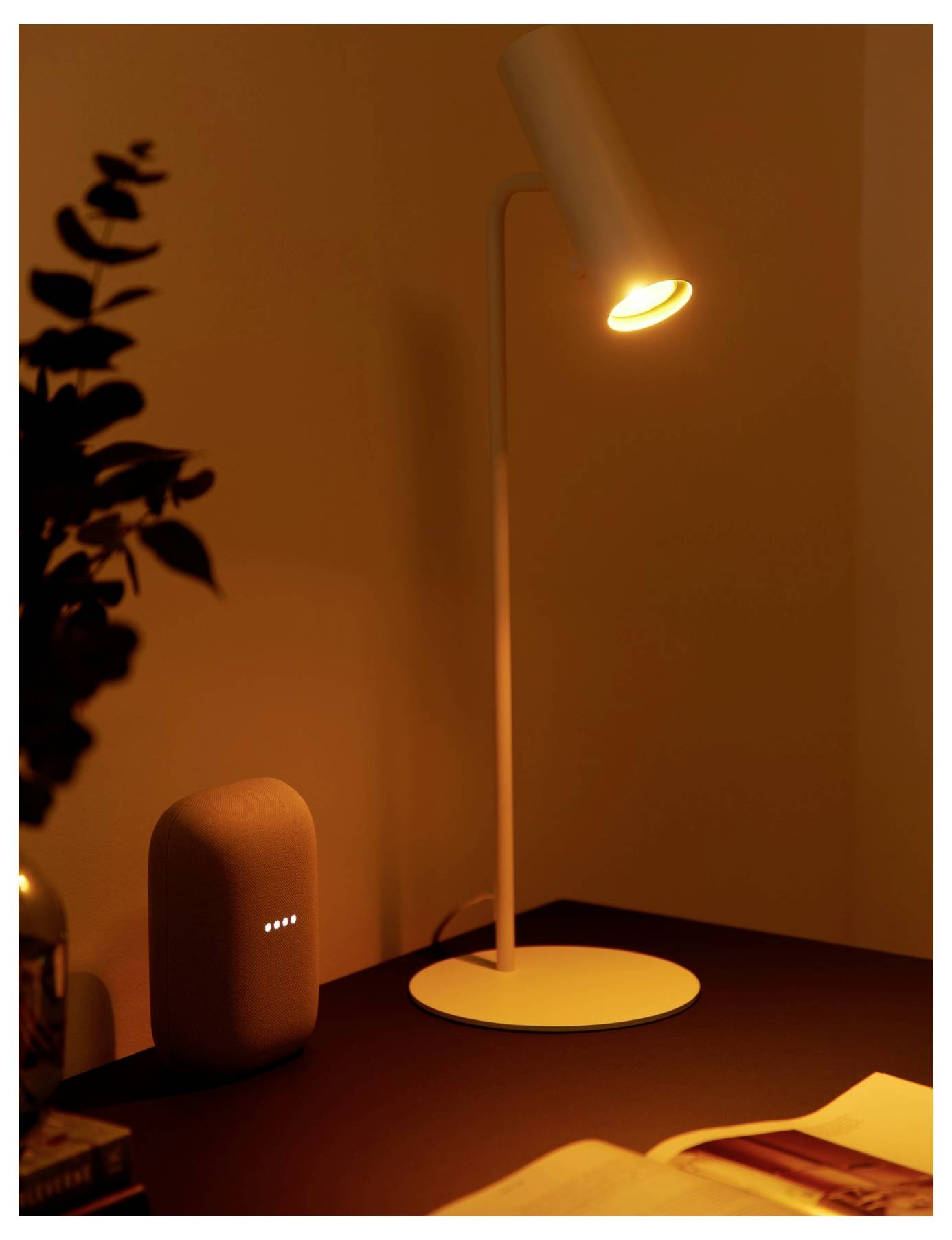 Une lampe de bureau émet une lumière chaude à côté d'un haut-parleur intelligent, avec des livres ouverts sur la table, créant une atmosphère confortable et chaleureuse.