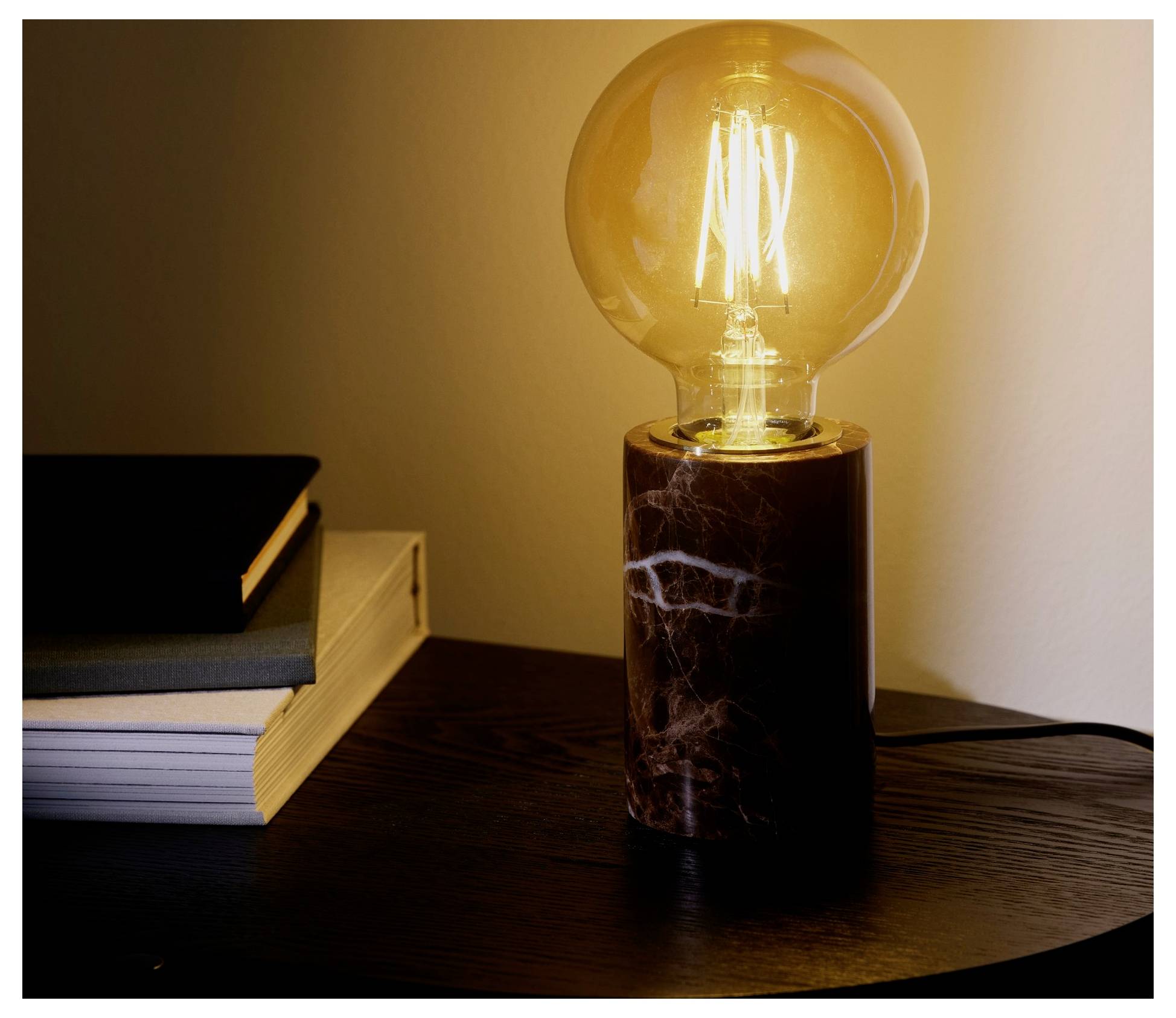 Une ampoule Edison brillante dans un socle en marbre à côté de livres empilés sur une table en bois foncé, créant une atmosphère chaleureuse et ambiante.