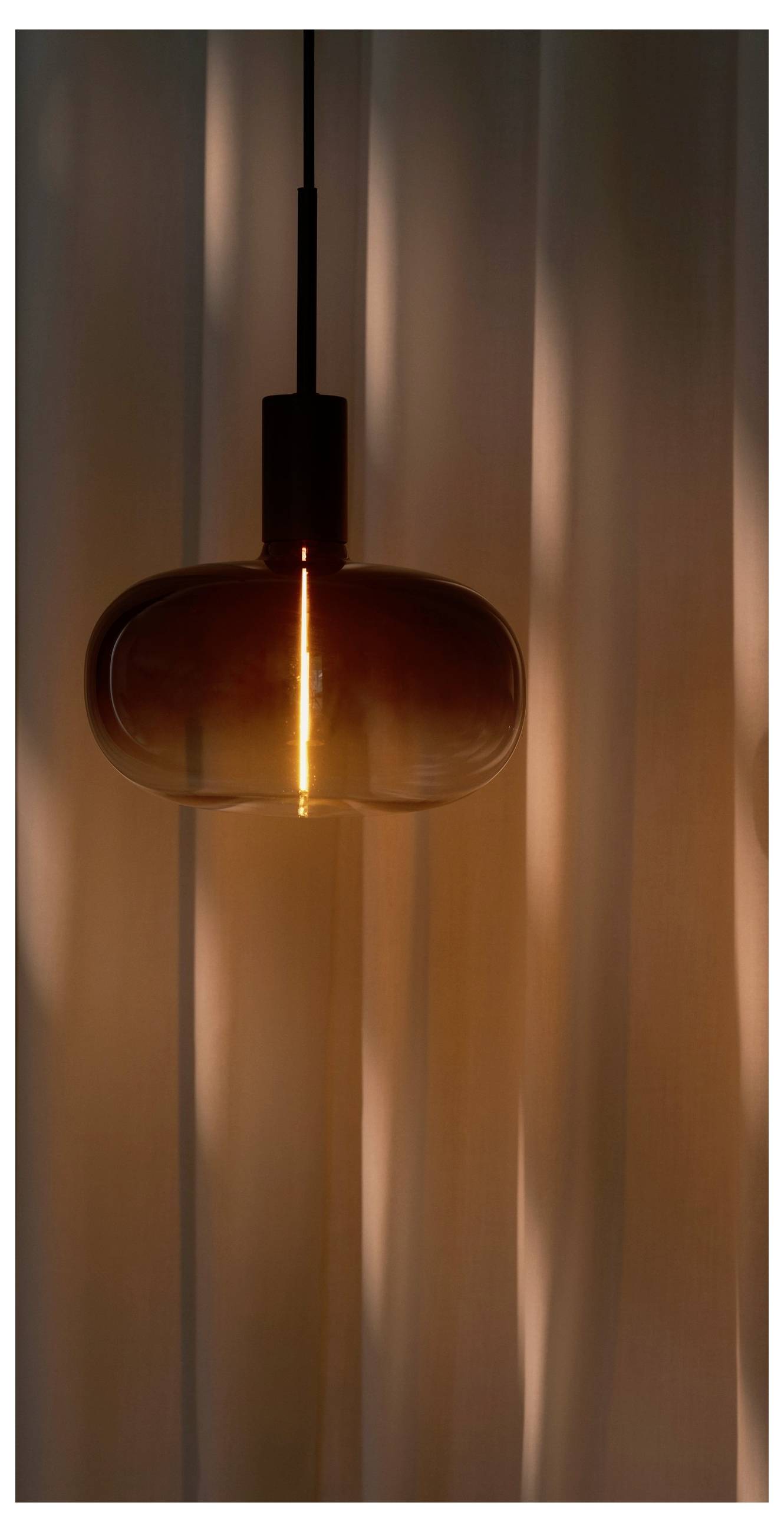 Une lampe suspendue chaleureuse et brillante avec un abat-jour en verre rond pend devant des rideaux doucement éclairés, créant une ambiance confortable et accueillante.