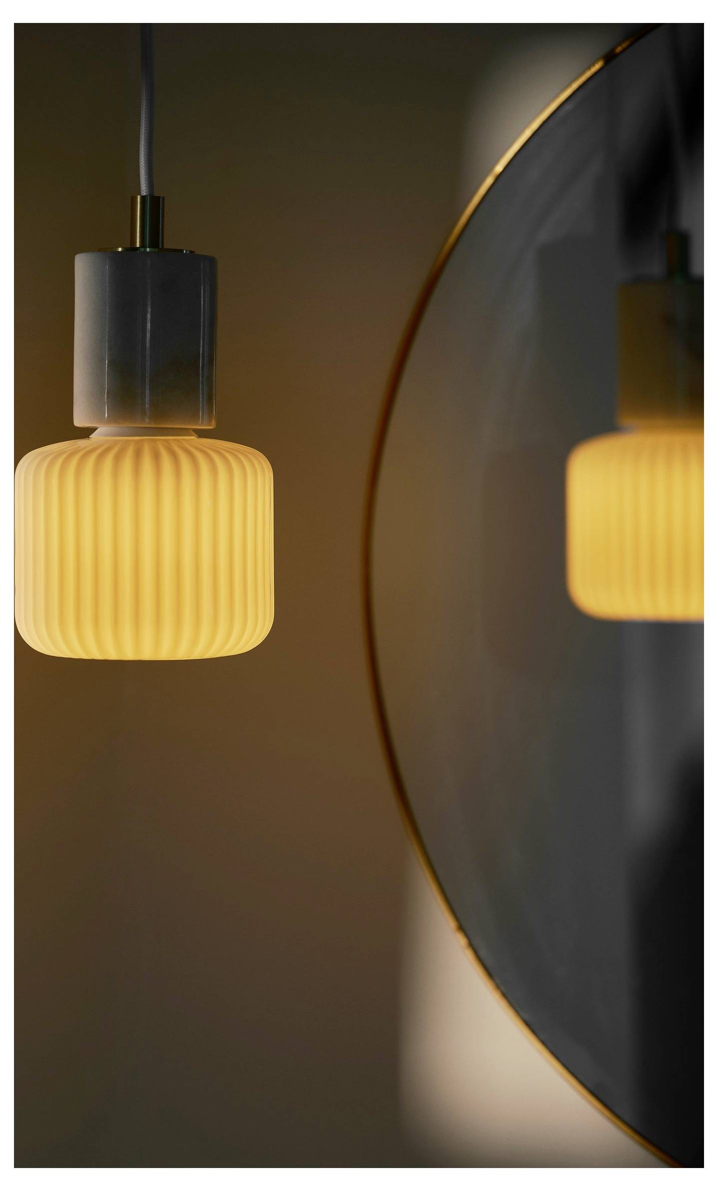 Un luminaire suspendu en verre nervuré unique émettant une lueur chaleureuse se reflète dans un miroir adjacent, créant un effet visuel symétrique.