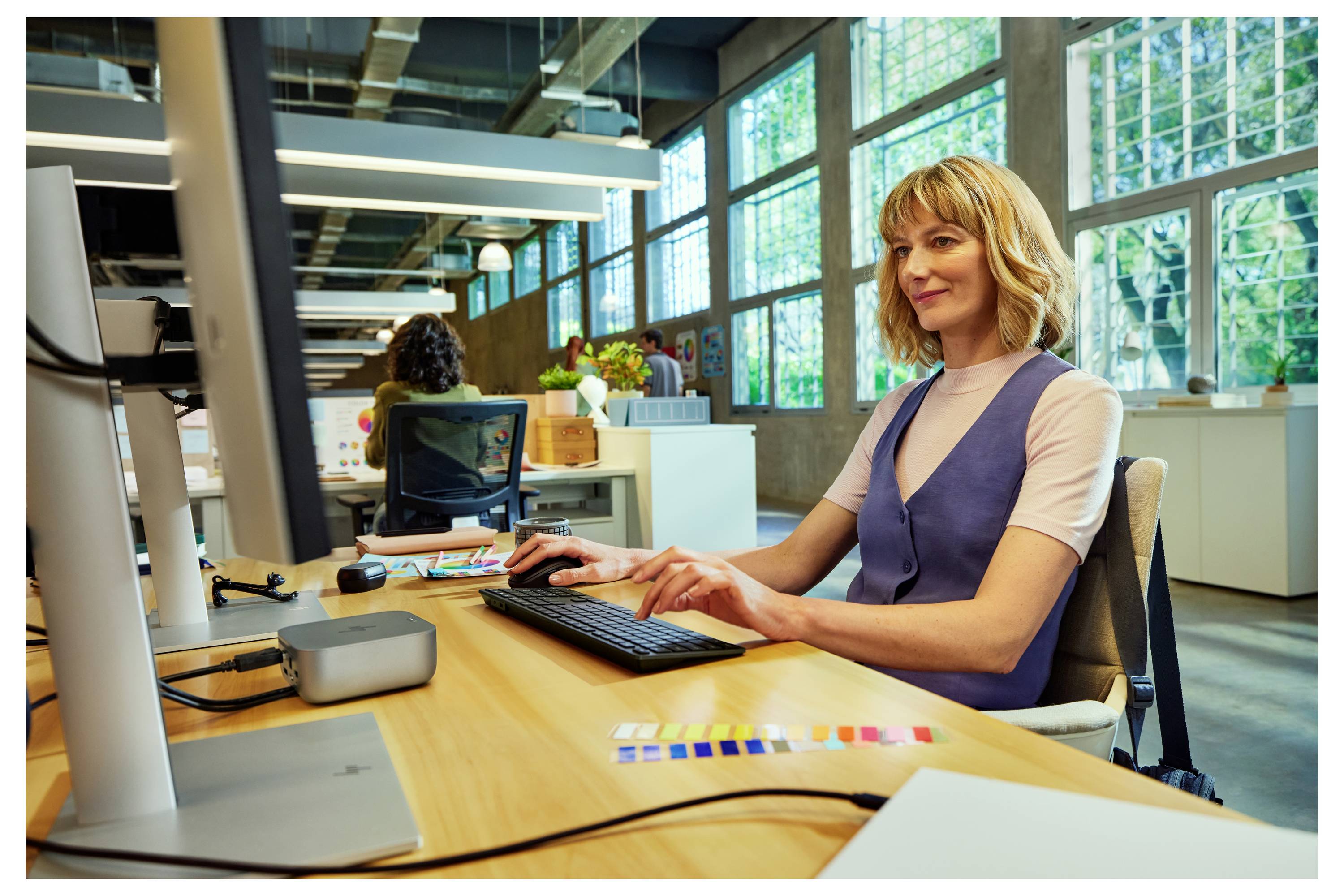 Une femme travaillant à un ordinateur dans un bureau moderne et bien éclairé avec de grandes fenêtres. Elle tape sur un clavier, l'air concentrée.