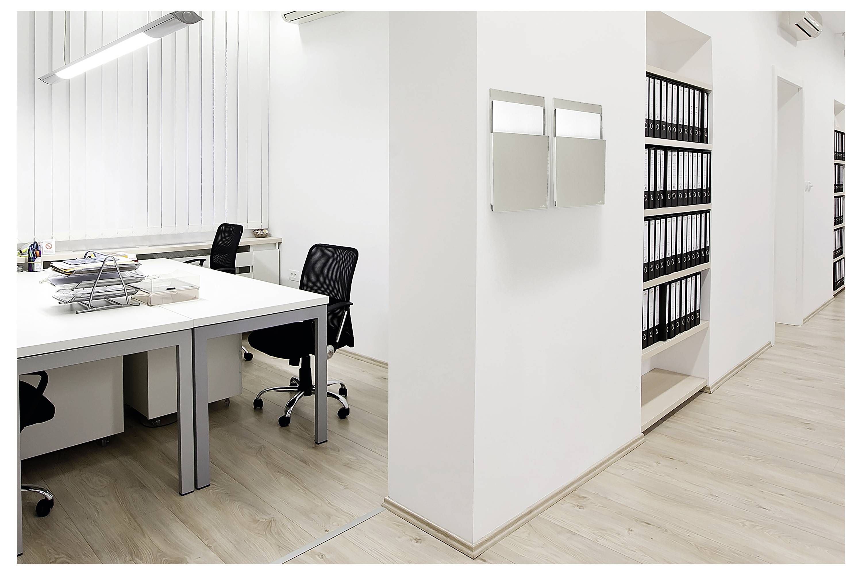 Espace de bureau moderne avec deux bureaux et des chaises, murs blancs, une pile de documents sur un bureau et des étagères remplies de classeurs noirs.