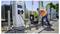 Un travailleur portant un gilet de sécurité connecte un câble à une borne de recharge pour véhicules électriques à l'extérieur, avec des voitures et des arbres en arrière-plan.