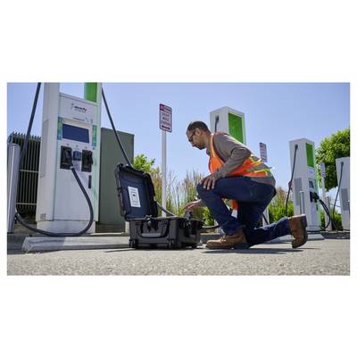 Une personne vêtue d'un gilet de sécurité s'agenouille à côté d'un boîtier d'équipement ouvert à une station de recharge pour véhicules électriques, examinant l'installation.